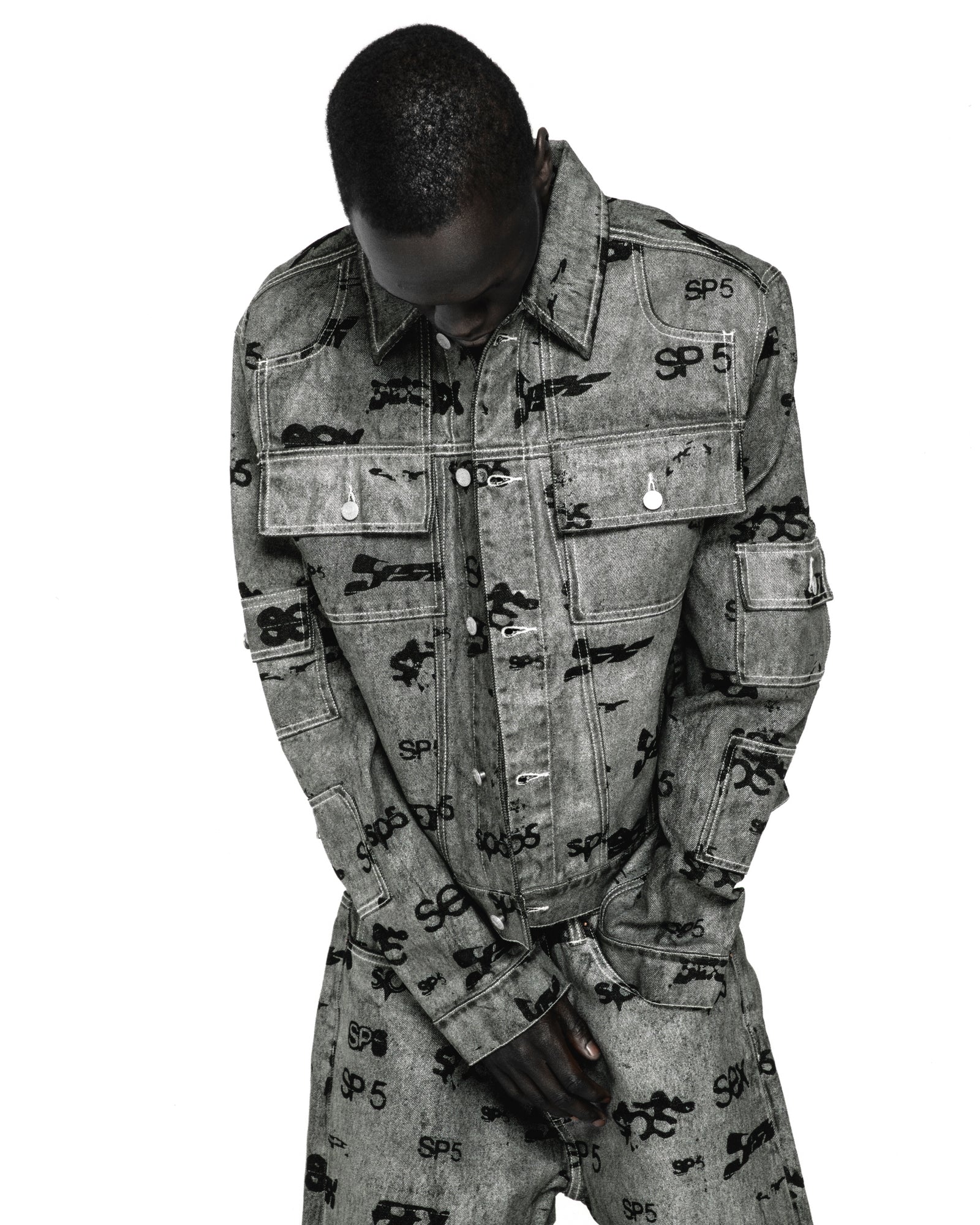 Gray denim trucker jacket with all-over black "SP5" print, silver buttons, and red contrast stitching.