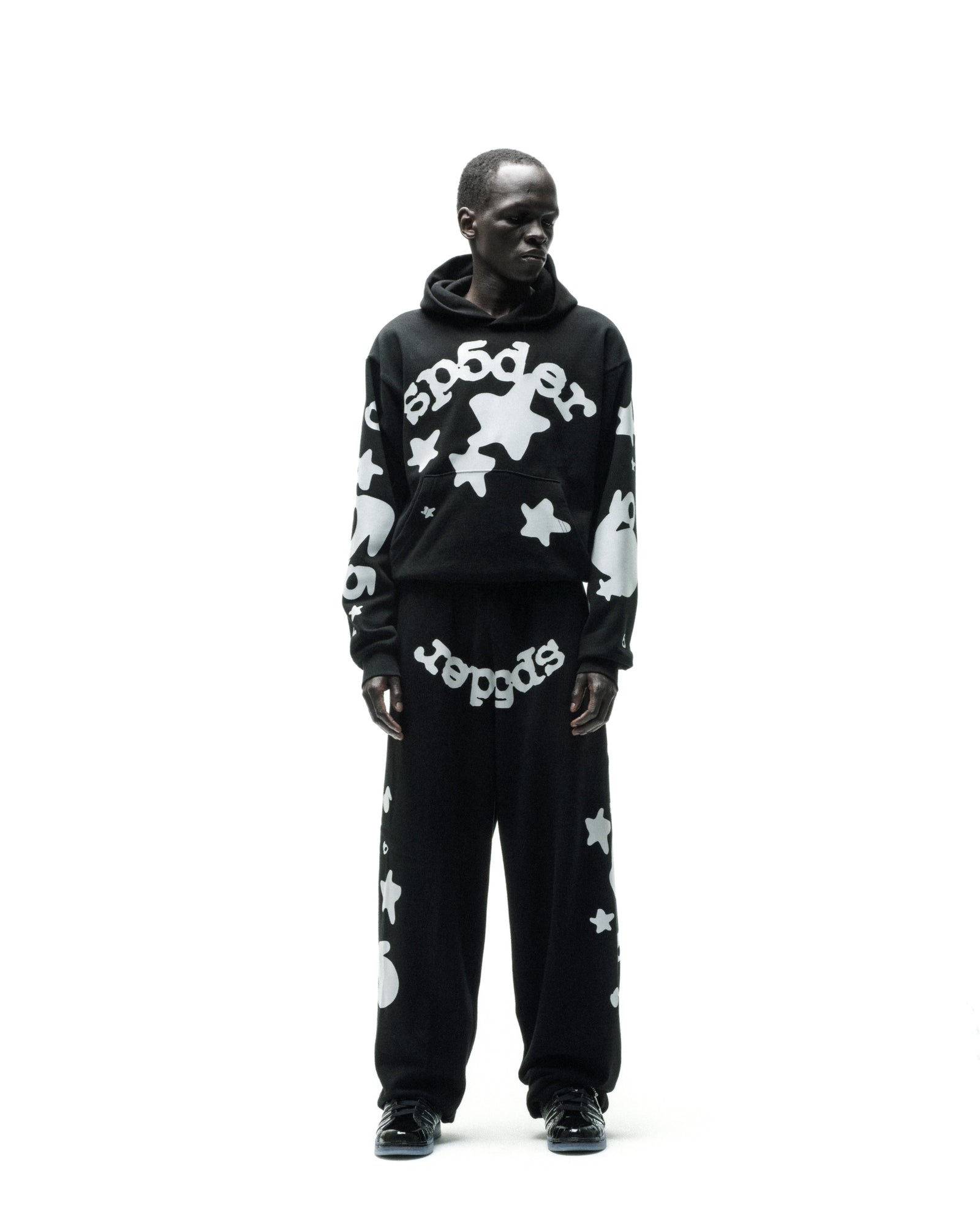 Man in black REFLECTIVE BELUGA HOODIE and sweatpants with white 'SP5DER' and star print.