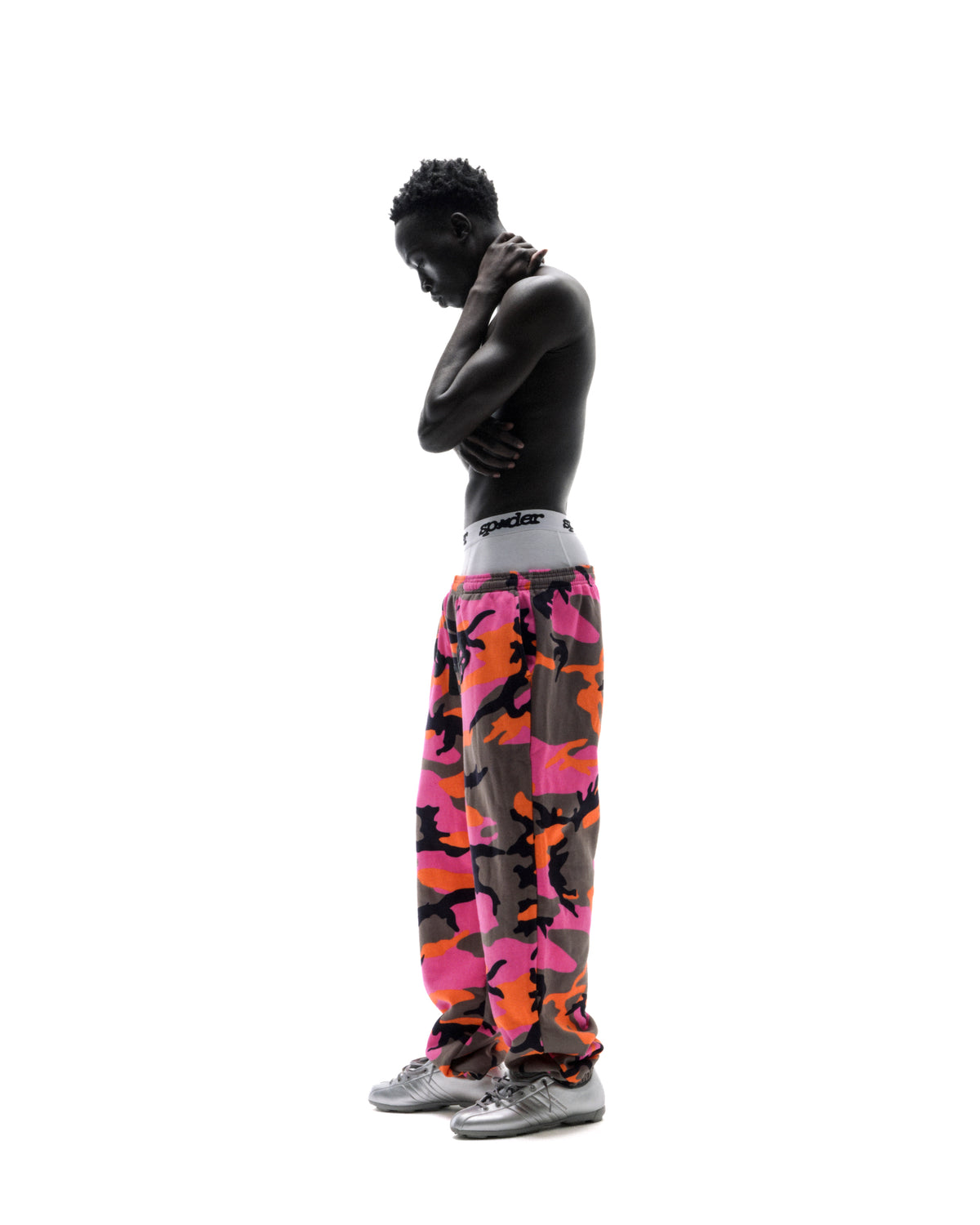 Shirtless model wearing pink and orange camouflage sweatpants and silver sneakers against a white background.