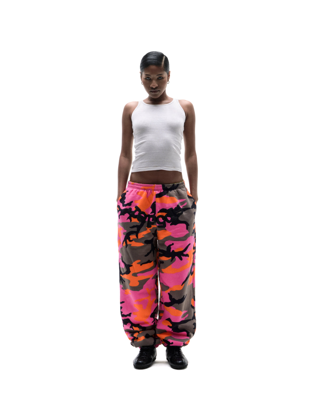 Model wearing a white ribbed tank top and vibrant pink and orange camouflage sweatpants against a white background.