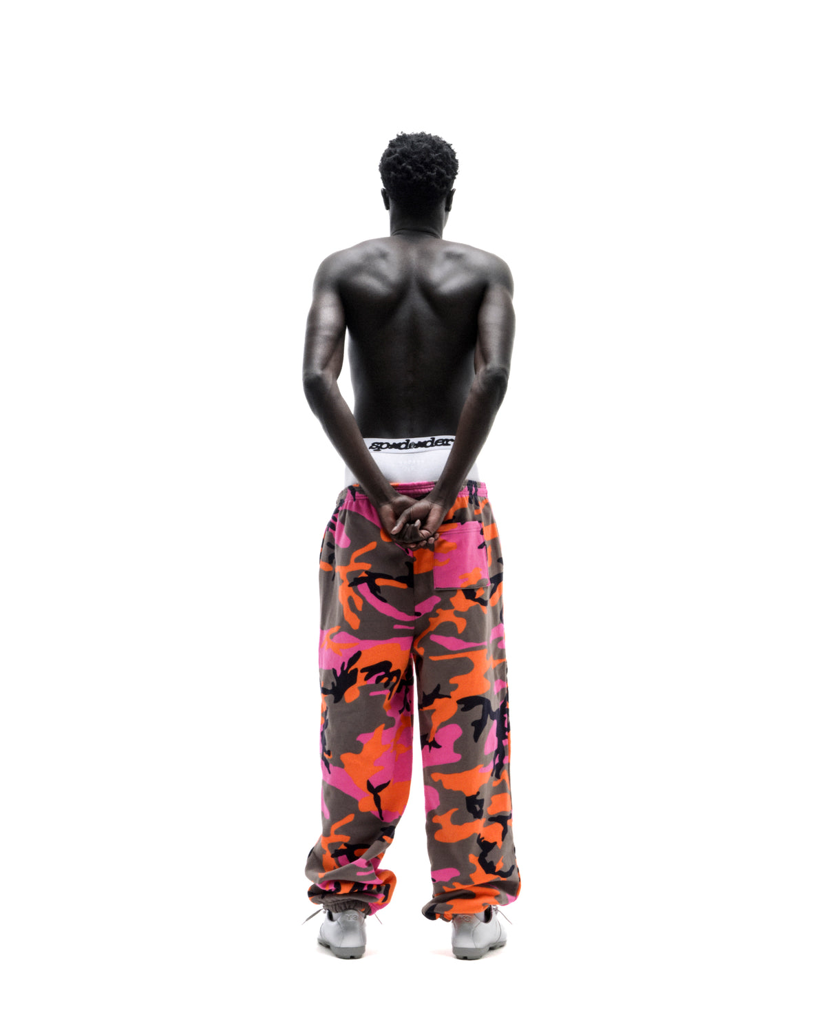Shirtless model standing with back turned, wearing pink and orange camouflage sweatpants and white sneakers.