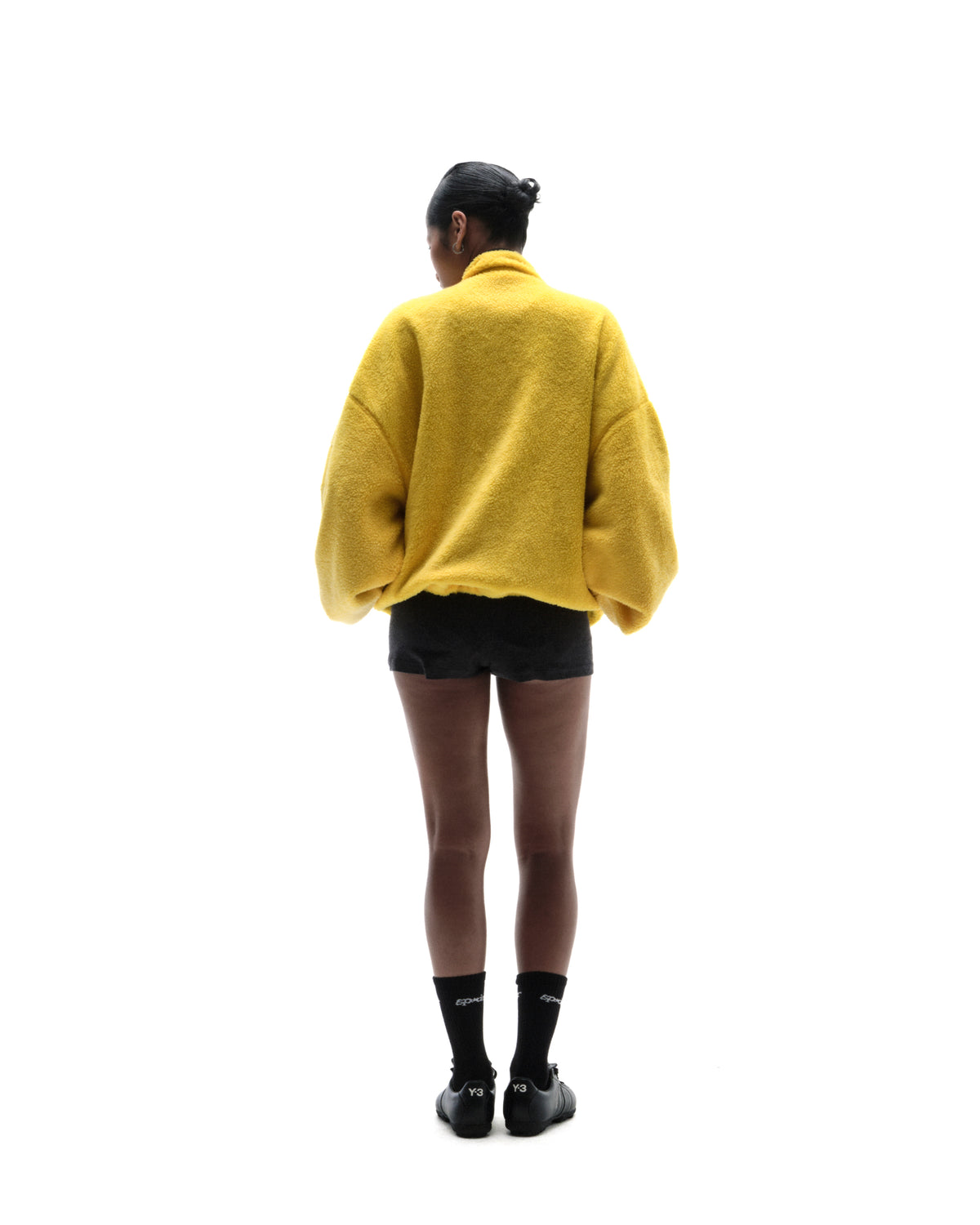 Model seen from behind wearing a bright yellow fleece jacket, black shorts, and black athletic sneakers with socks.