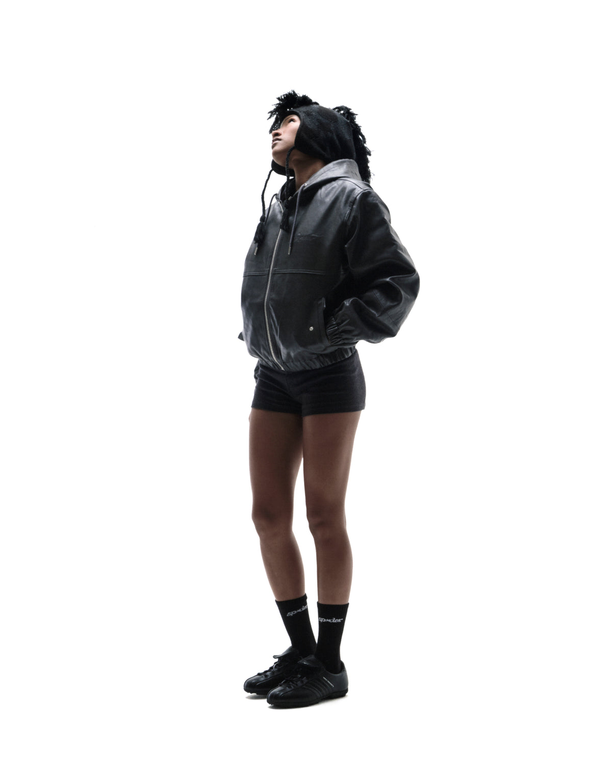 Model wearing a black leather bomber jacket, shorts, and a textured knit hat, standing against a white background.