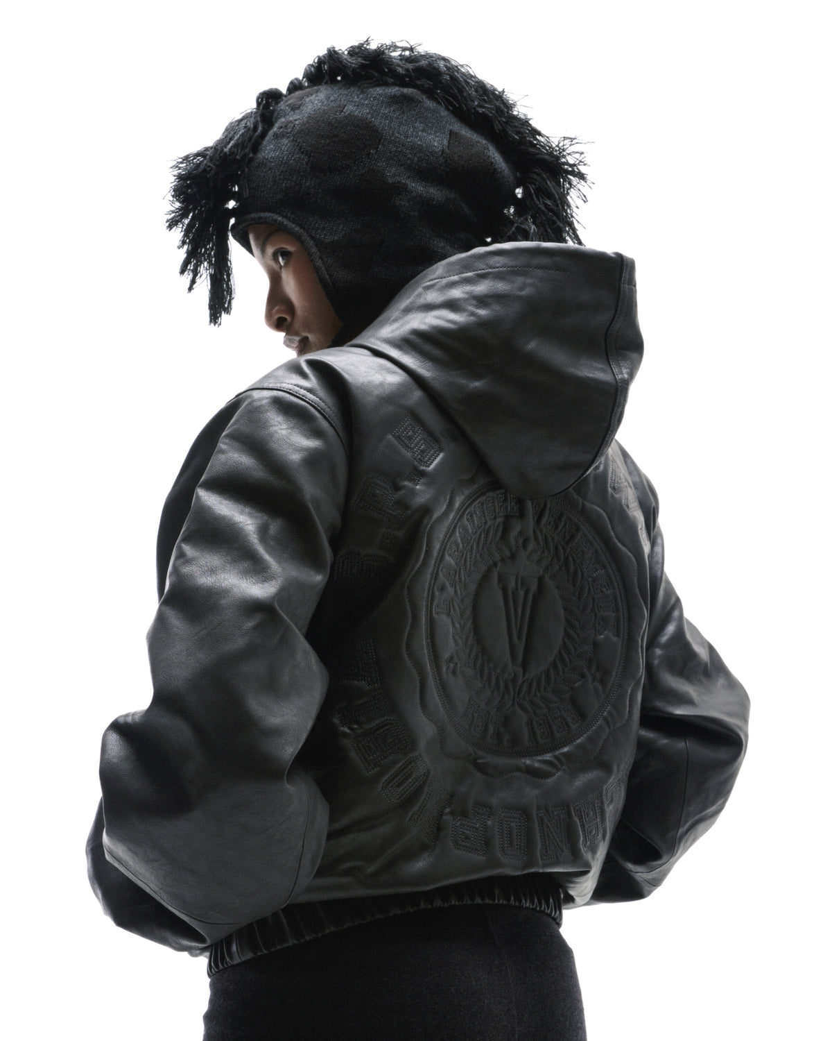 A model wearing a black leather hooded jacket with a large circular embossed logo on the back and a textured knit hat.