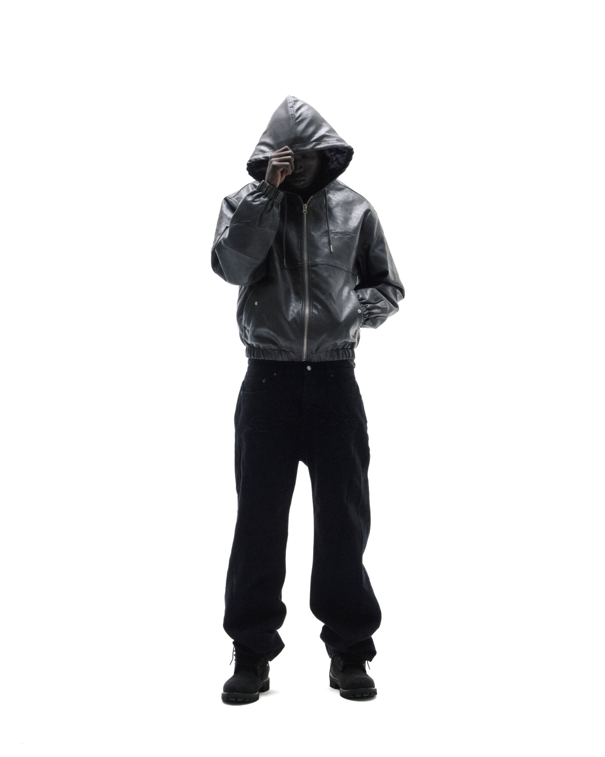 Model wearing a black leather hooded jacket and loose-fit dark trousers against a white background.
