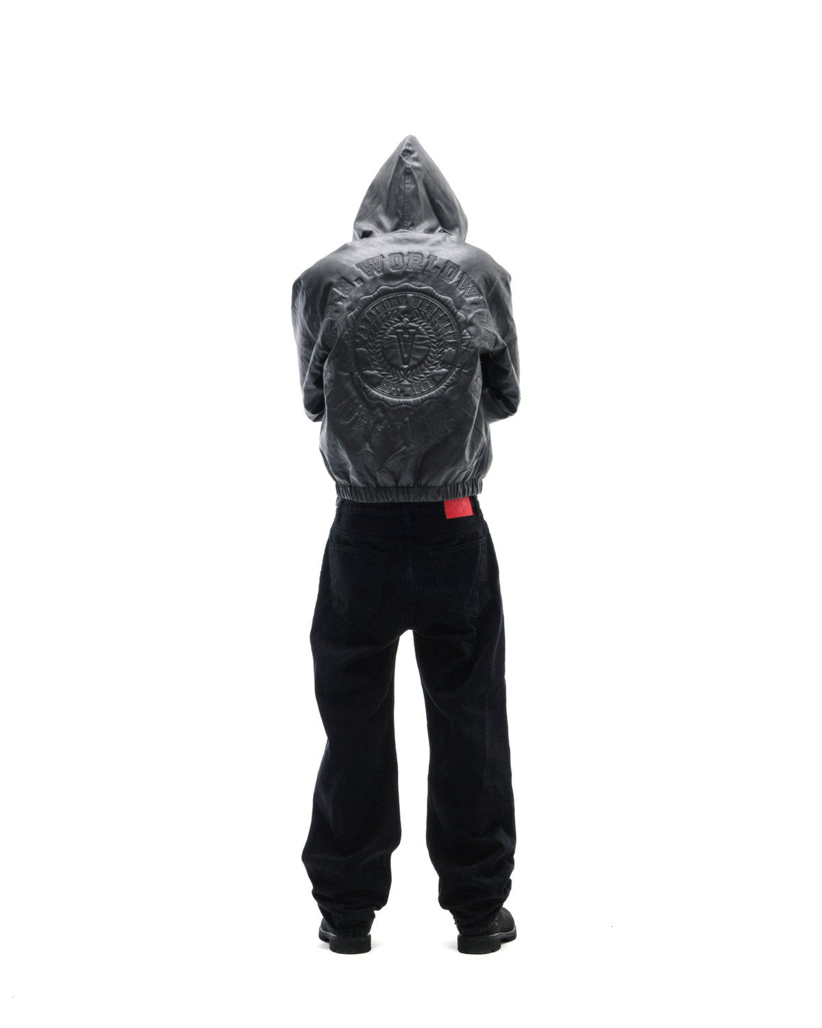 Model wearing a black leather hooded jacket with embossed back detailing and loose-fit black jeans against white.