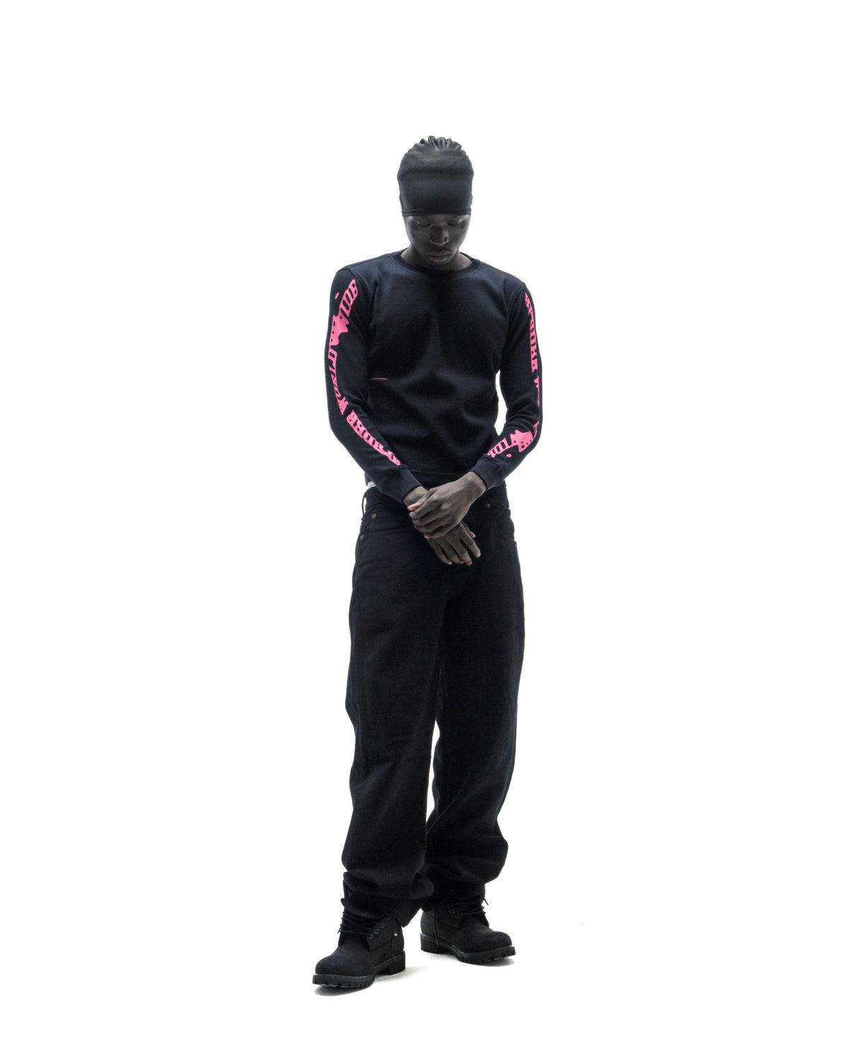 Model wearing a black long-sleeve shirt with pink text on the sleeves, black baggy jeans, and a black skull cap.