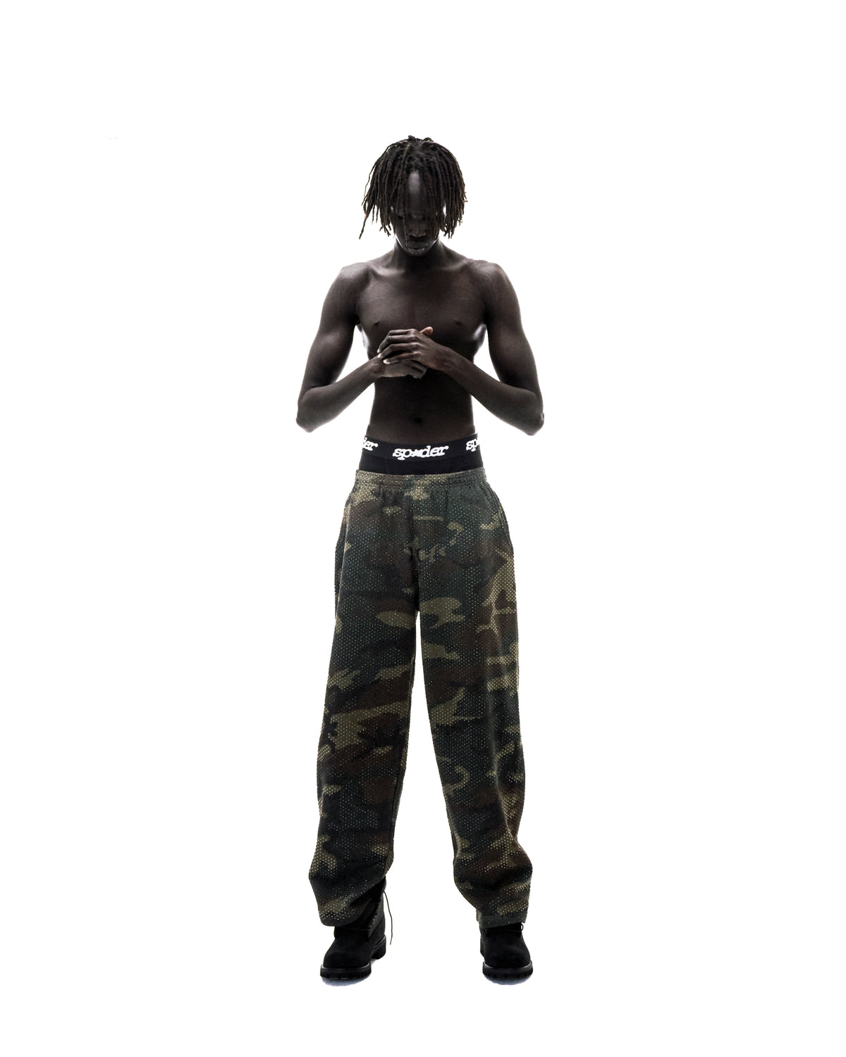 Model with dreadlocks wearing camouflage mesh pants and black Sp5der branded underwear against a white background.