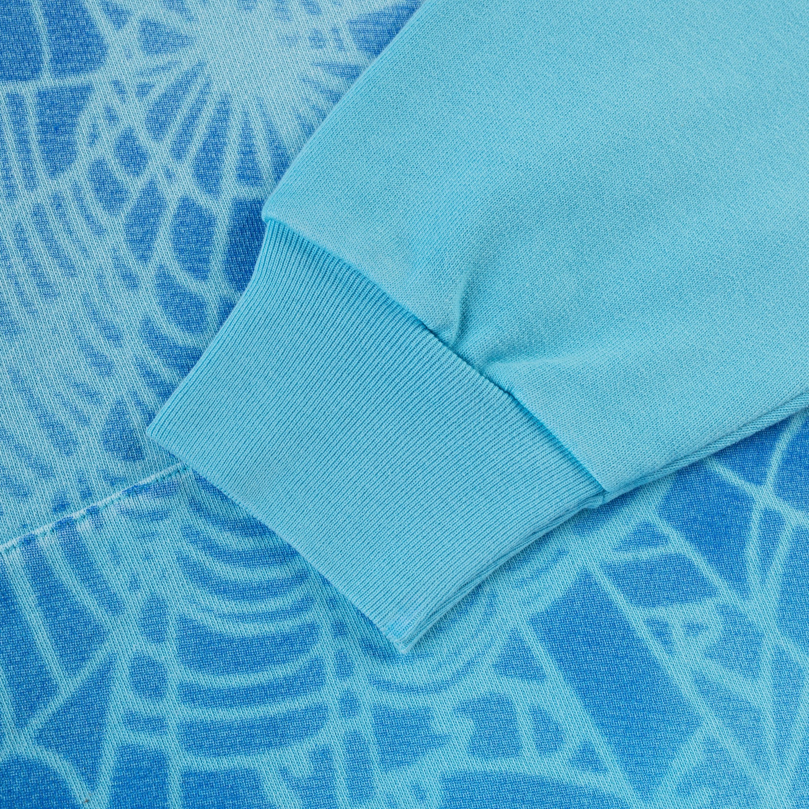 Close-up of a light blue cotton fleece hoodie with a darker blue spray web V2 print and ribbed cuff.