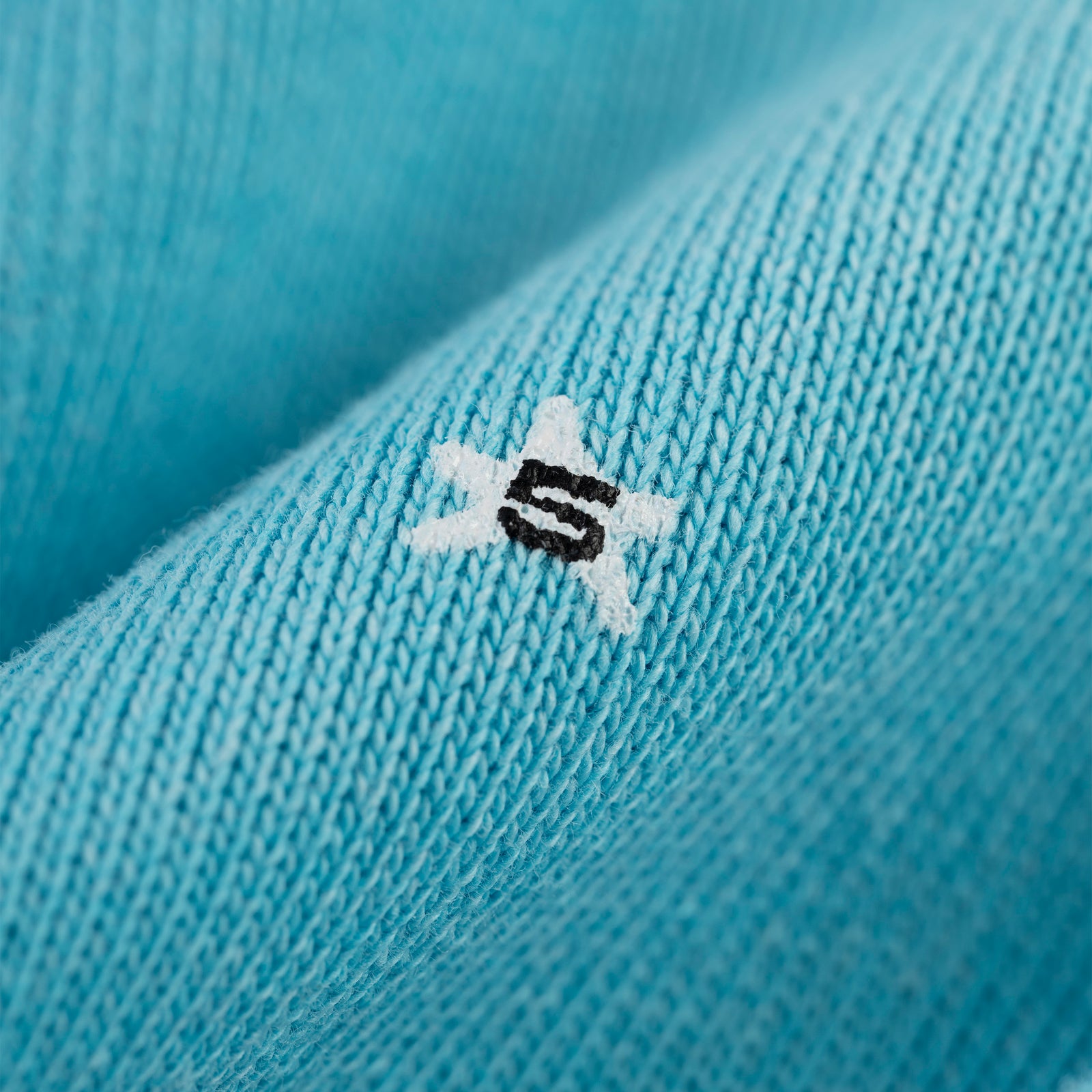 Close-up of a turquoise cotton fleece hoodie with a white star and black "S" puff print.