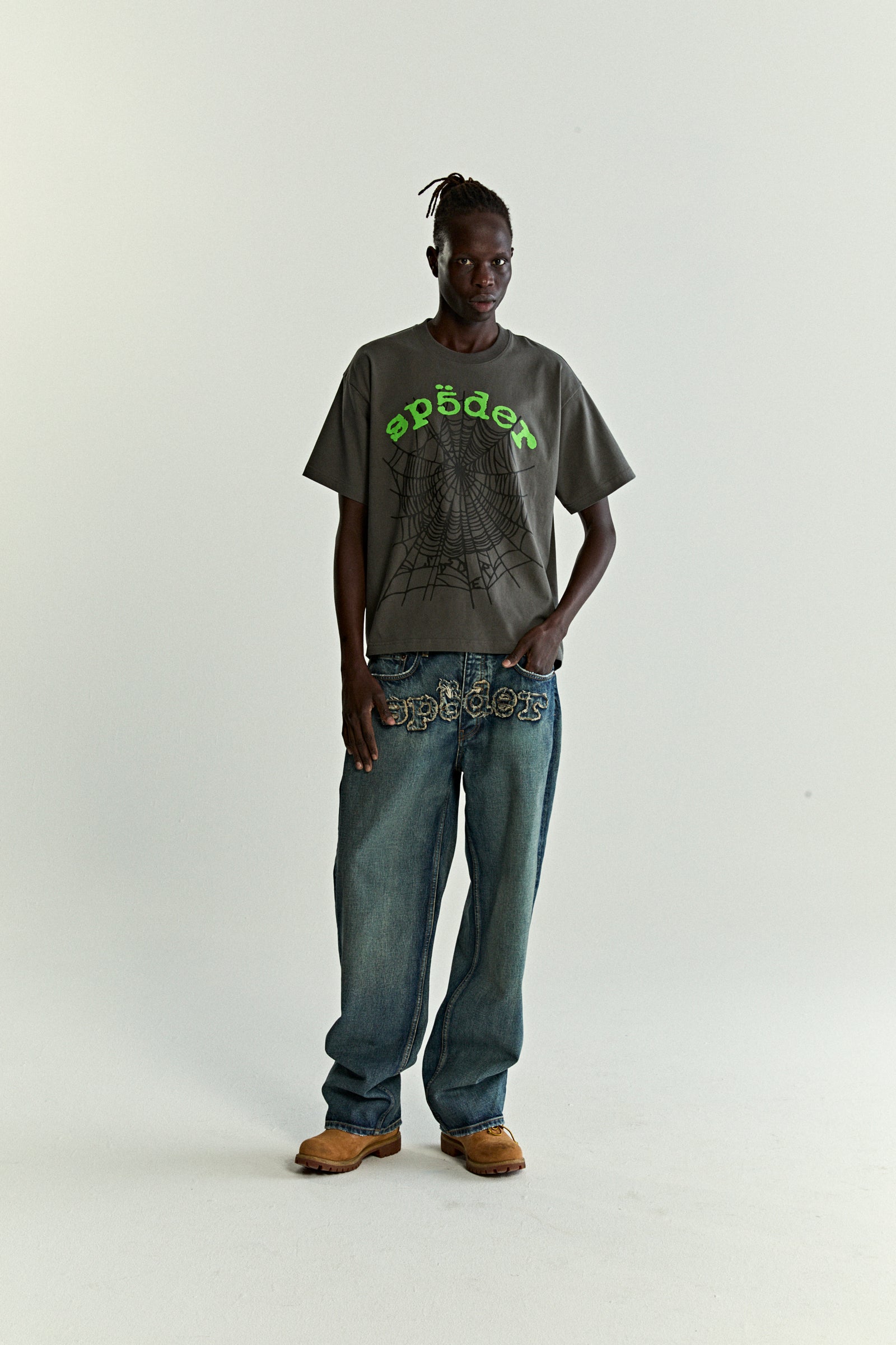 Model wearing an OG WEB V2 TEE, a gray cotton tee with a green spider logo and web print.