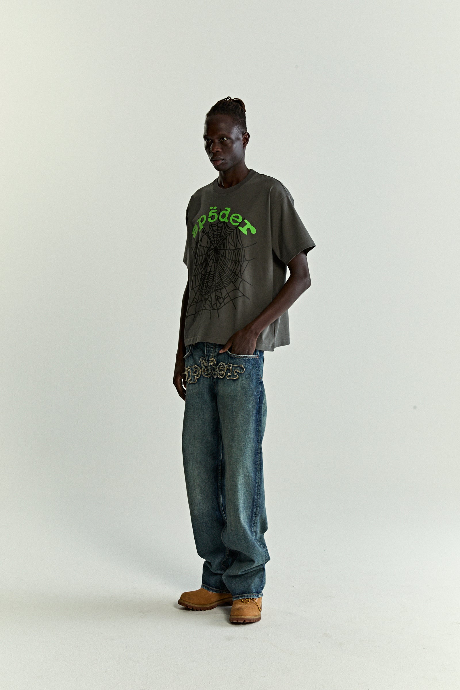 Man wearing a grey OG WEB V2 TEE with green puff print spiderweb design and baggy jeans.