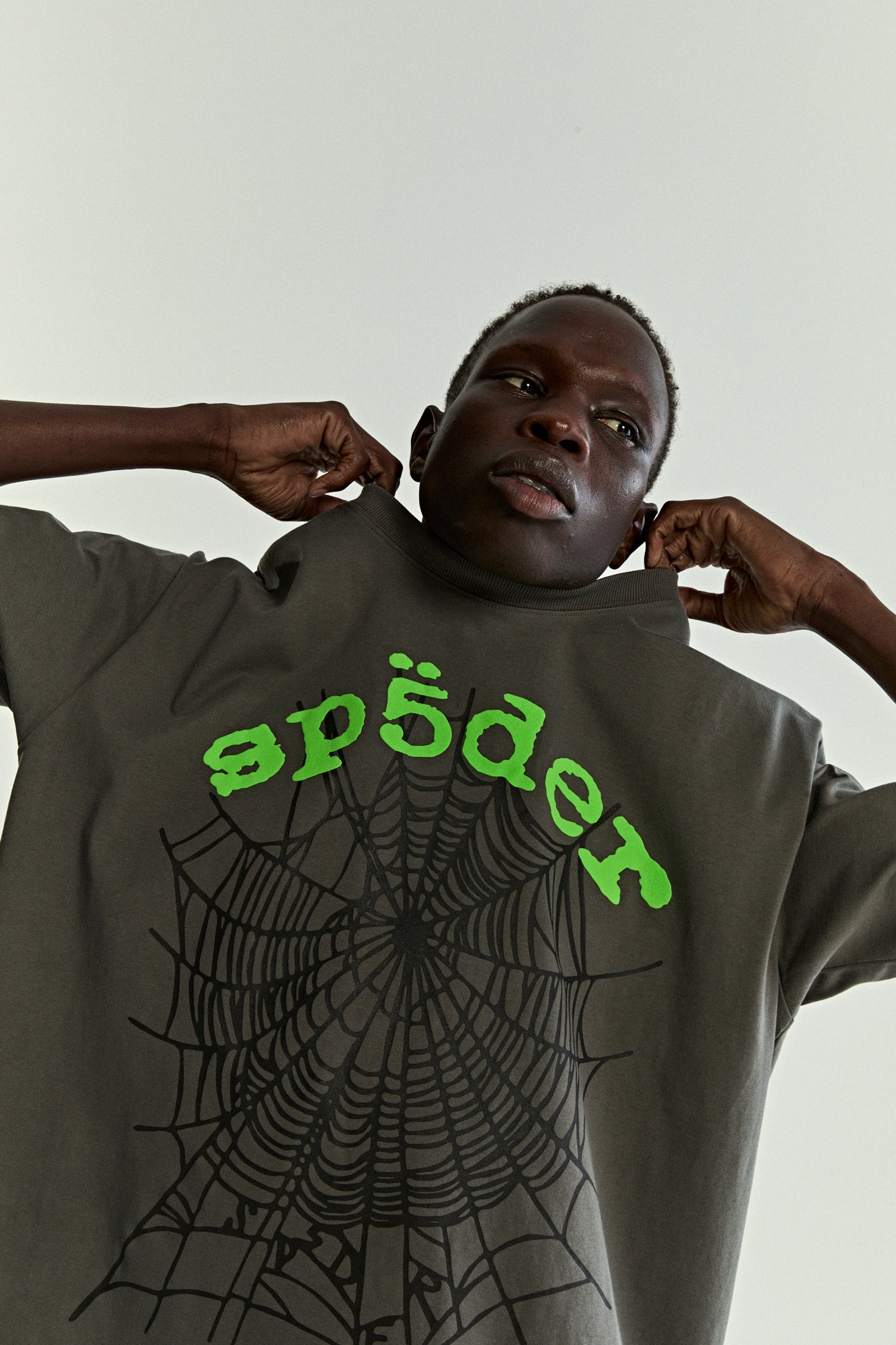 Man wearing a dark grey OG WEB V2 TEE with bright green "sp5der" text and a black spiderweb graphic.