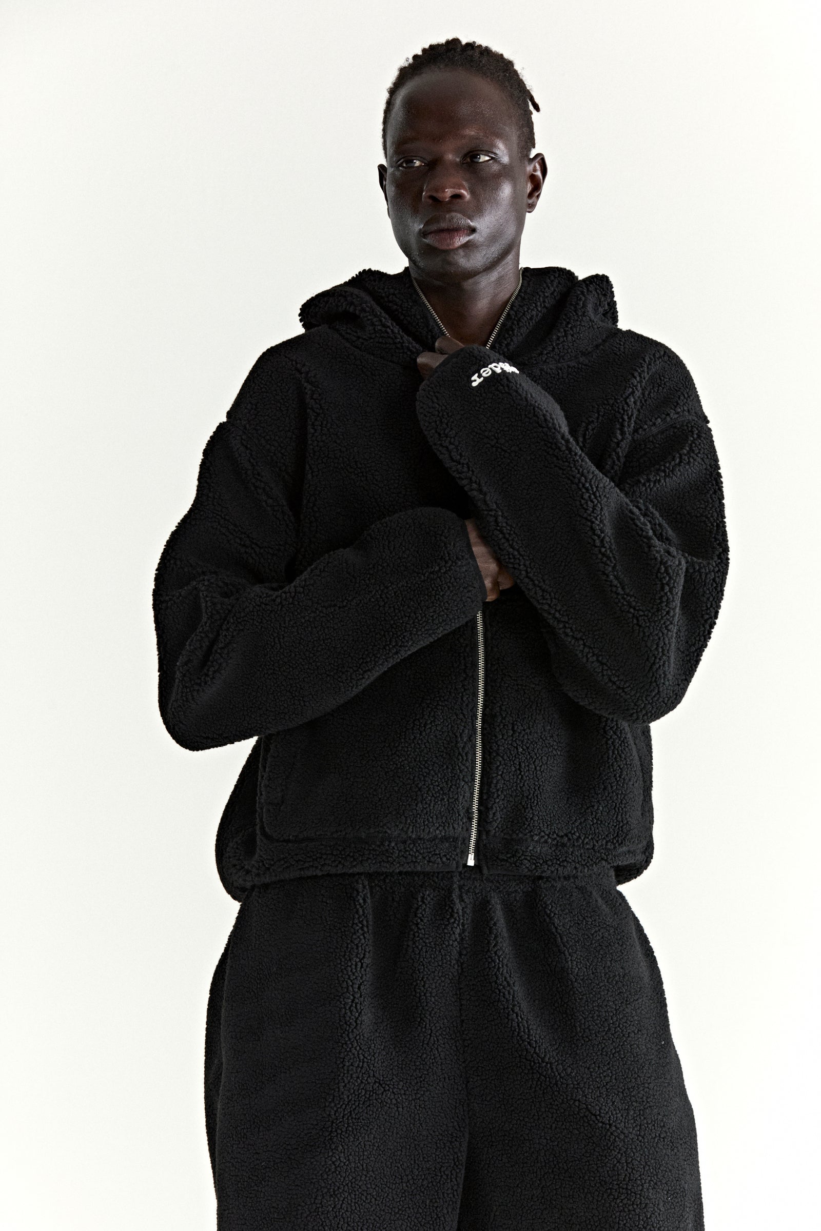 Black teddy fleece zip-up hooded jacket with rubberized logo patch on wrist, worn by a model.