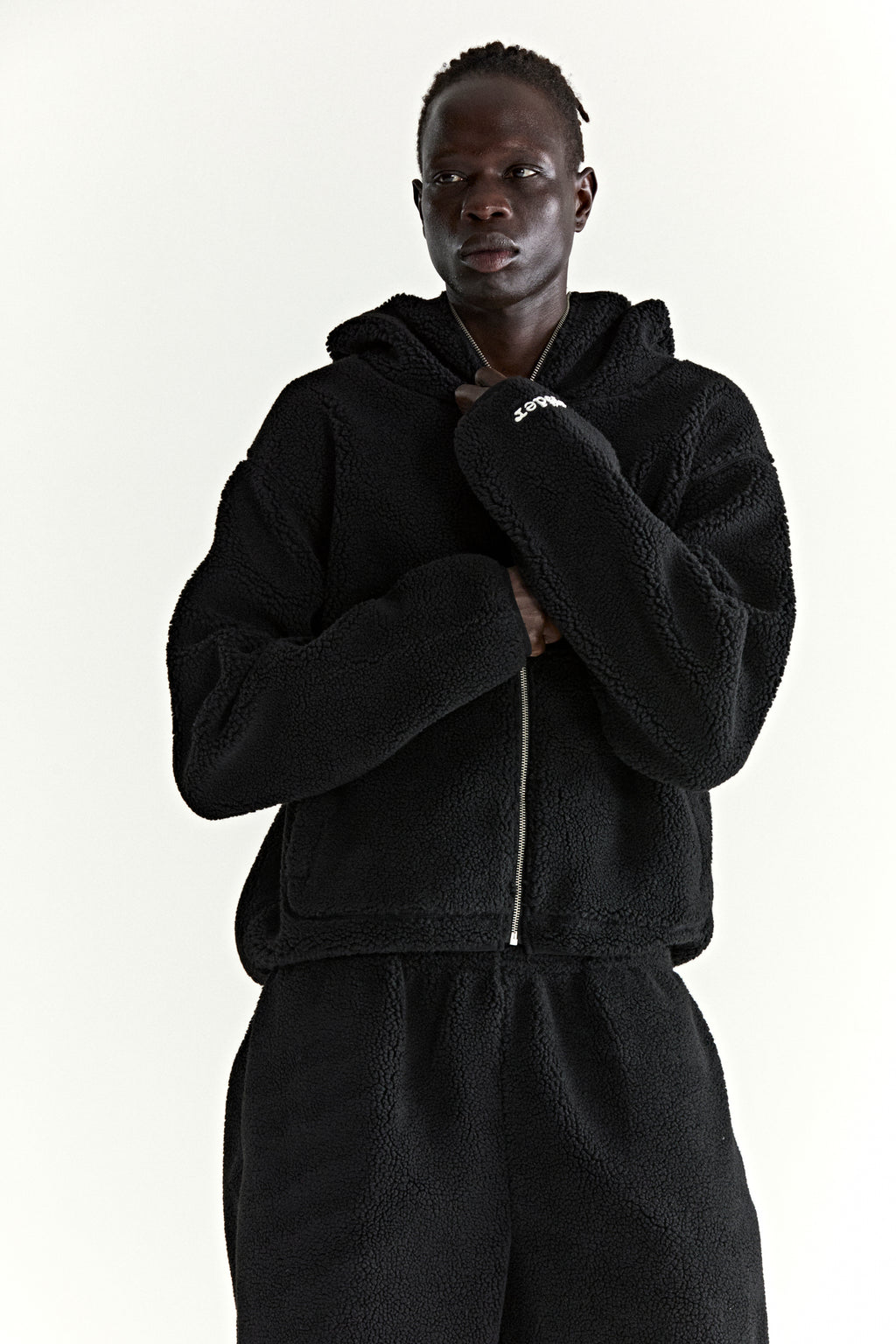 Black teddy fleece zip-up hooded jacket with rubberized logo patch on wrist, worn by a model.