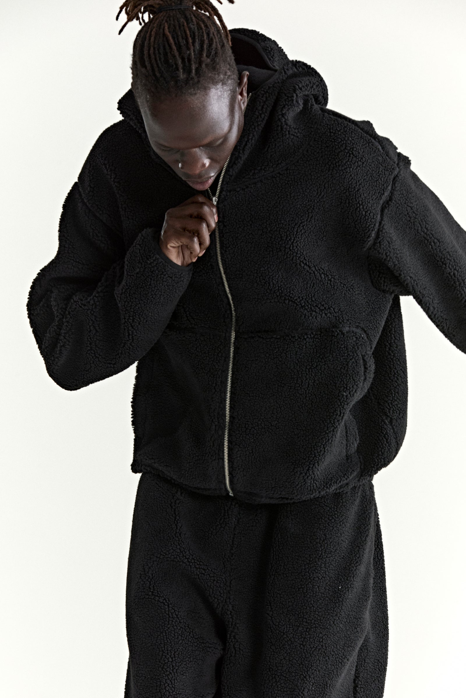 Black teddy fleece zip-up hooded jacket with welt pockets, worn by a person zipping it up.