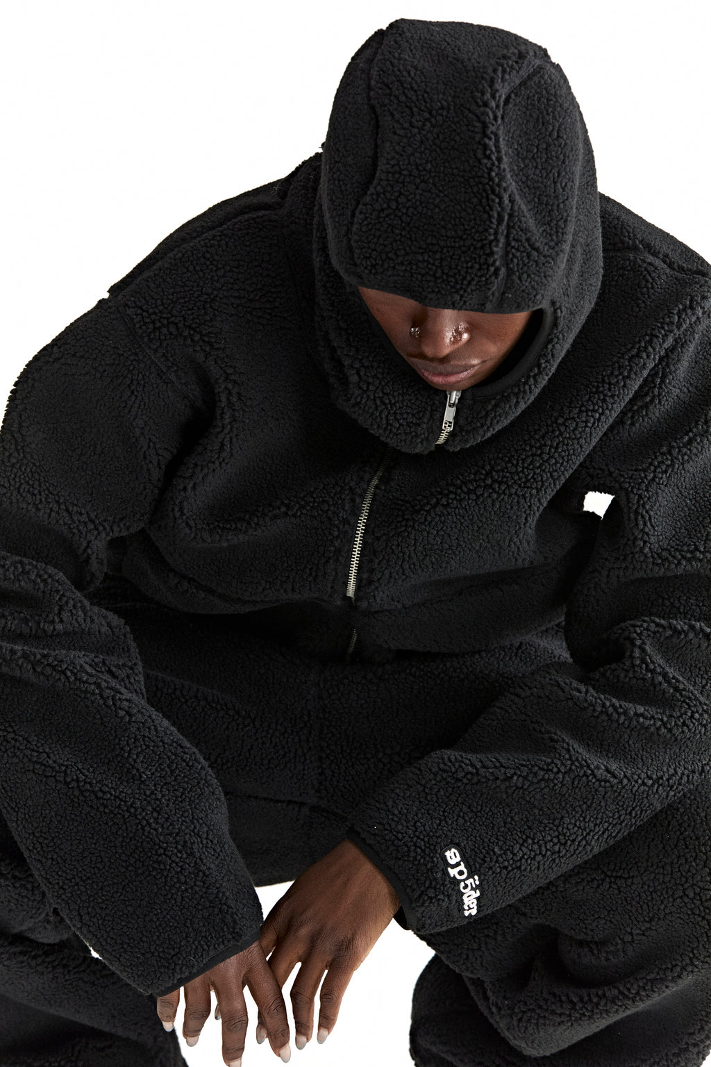 Black teddy fleece zip-up hooded jacket with rubberized logo patch on wrist, worn by a person.
