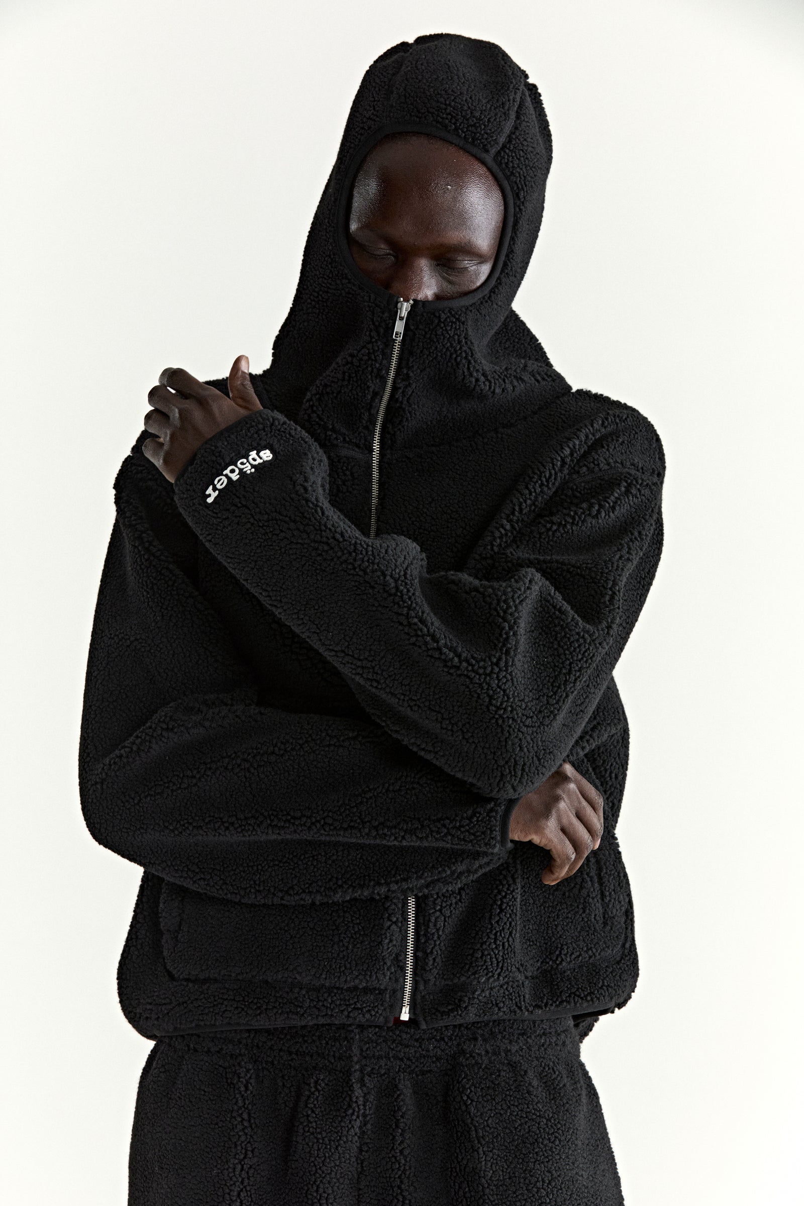 Black teddy fleece zip-up hooded jacket with rubberized logo patch on wrist and welt pockets.