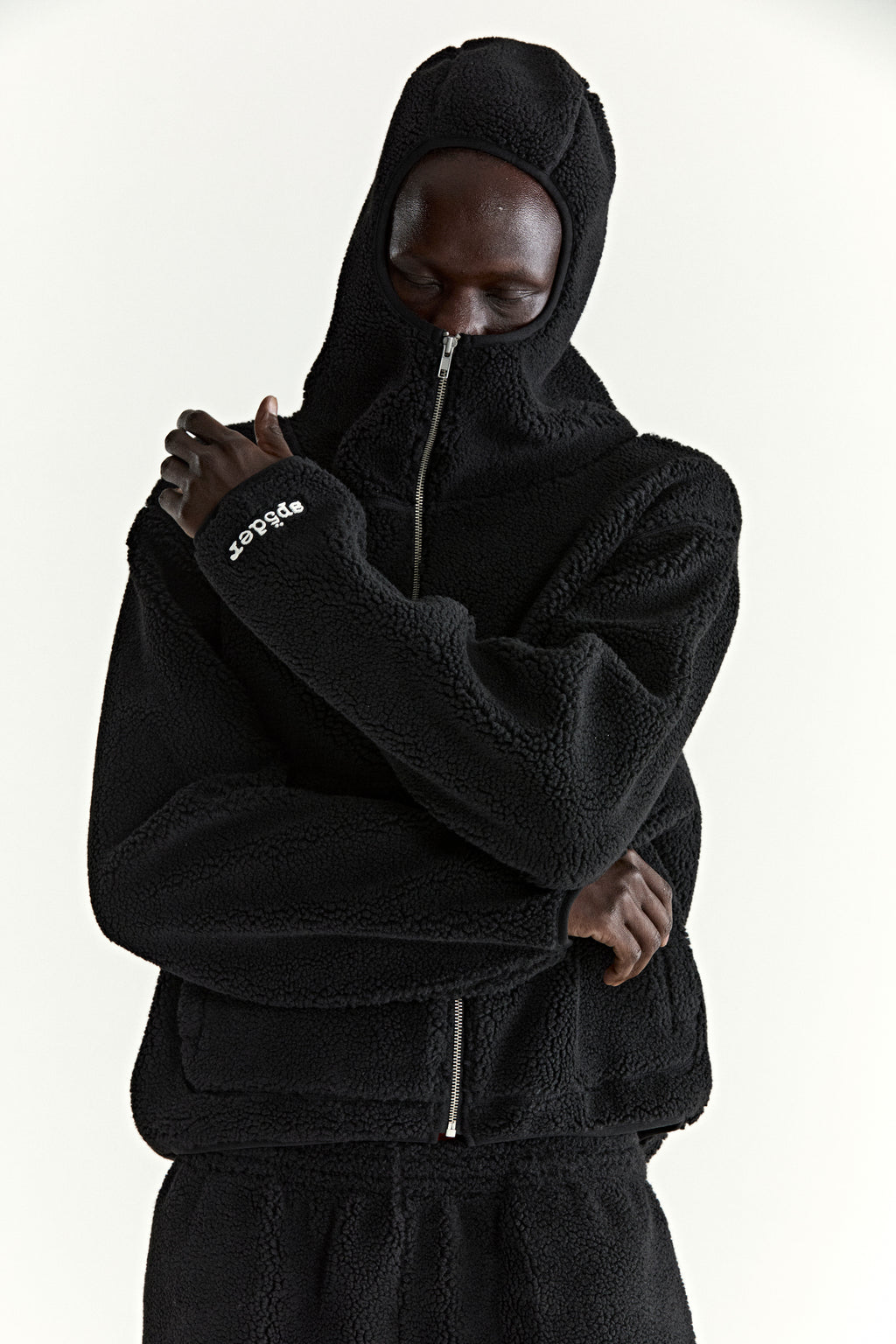 Black teddy fleece zip-up hooded jacket with rubberized logo patch on wrist and welt pockets.