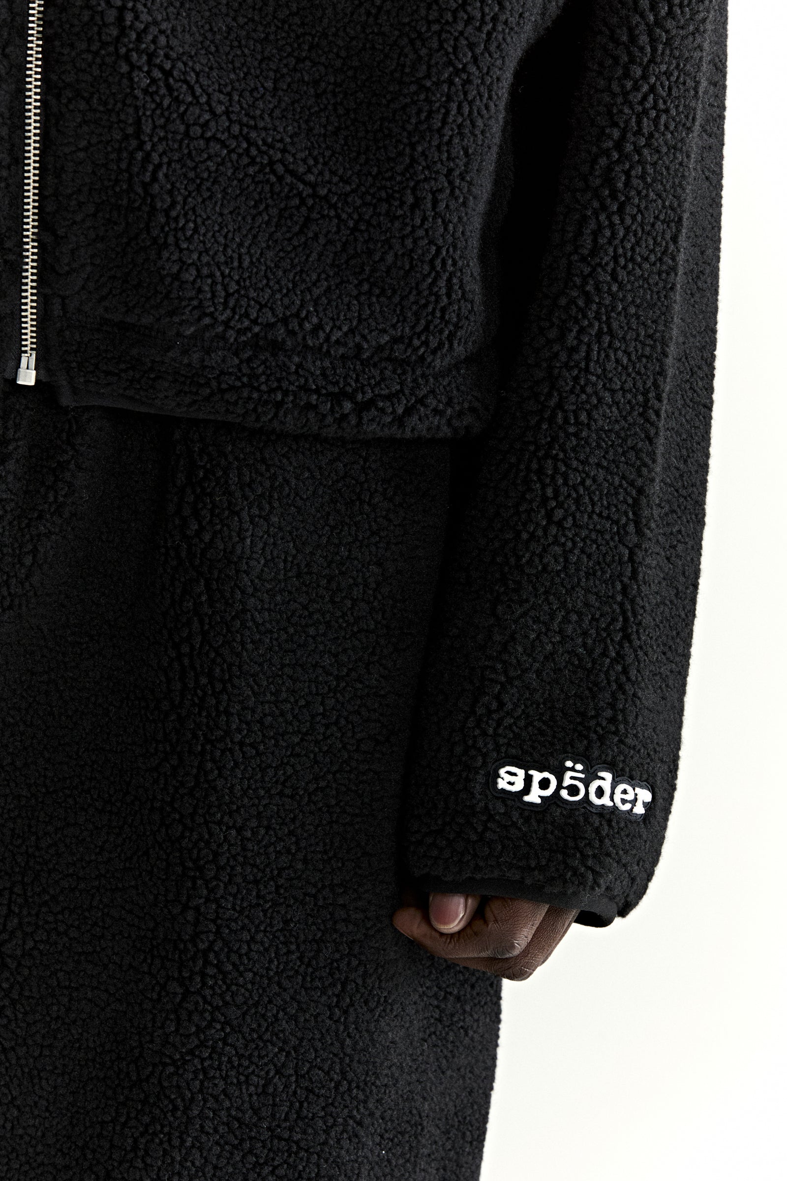 Black teddy fleece zip-up jacket with white "sp5der" logo on sleeve and silver zipper.