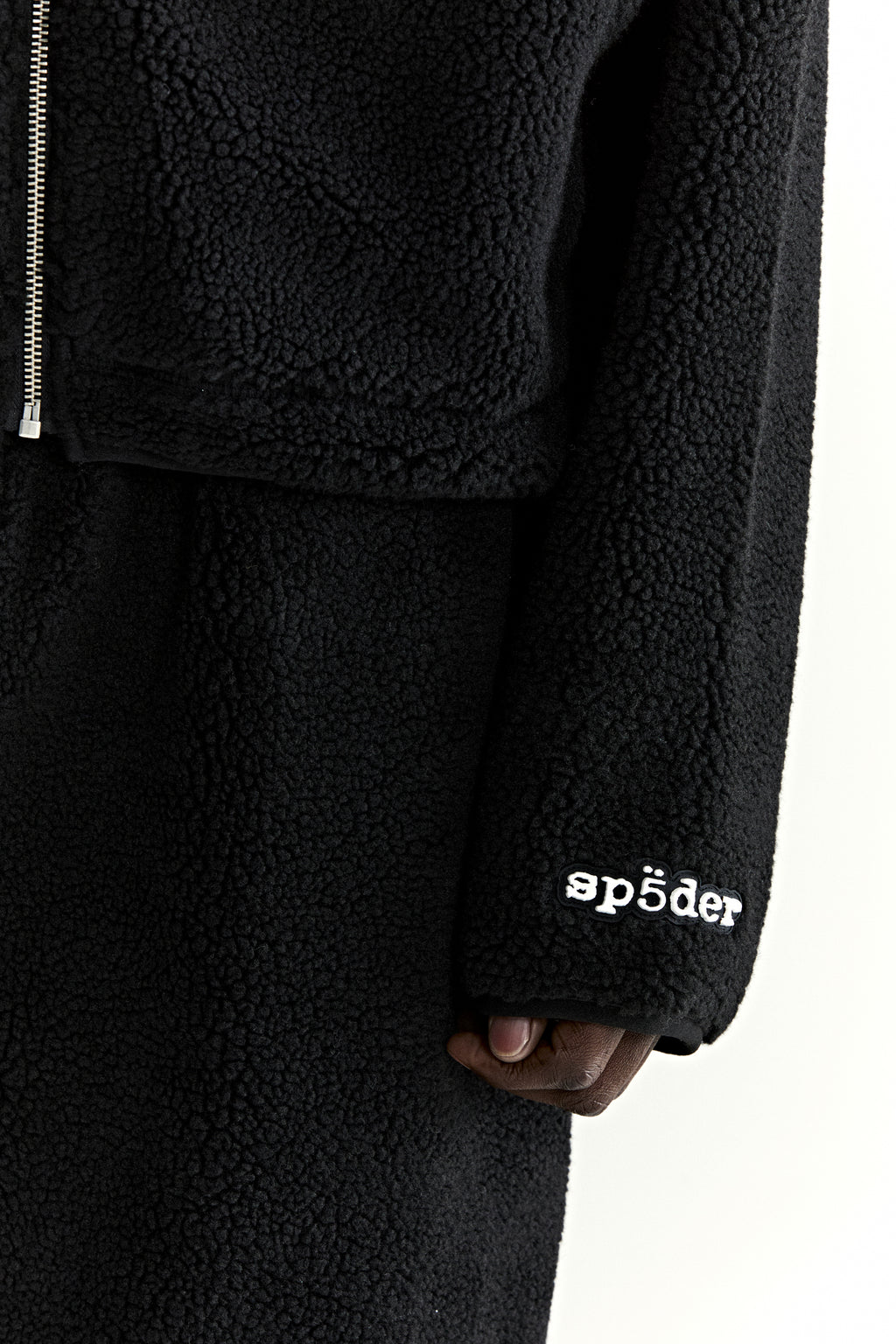 Black teddy fleece zip-up jacket with white "sp5der" logo on sleeve and silver zipper.