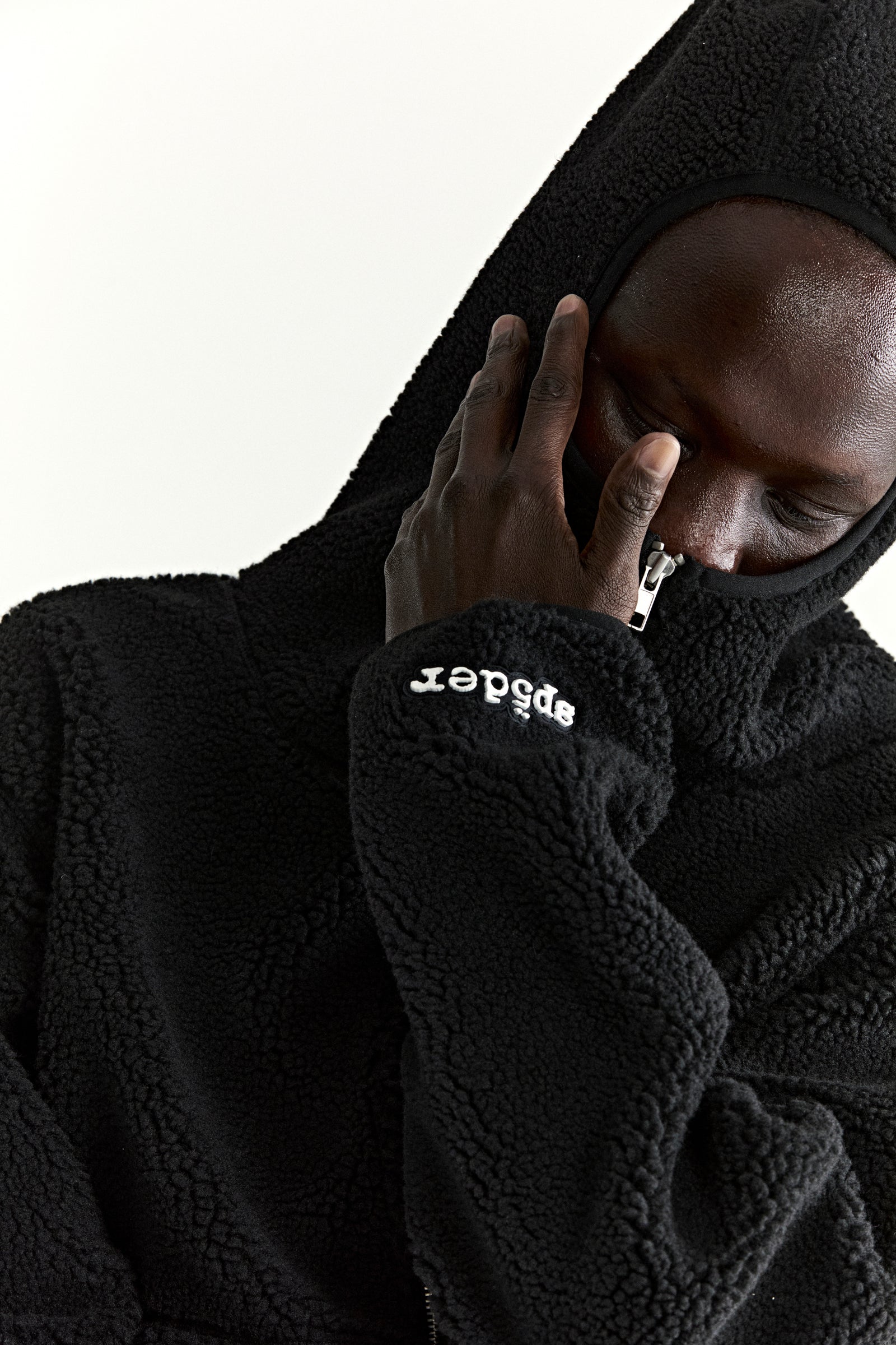 Black teddy fleece zip-up jacket with hood, rubberized logo patch on wrist, and welt pockets.