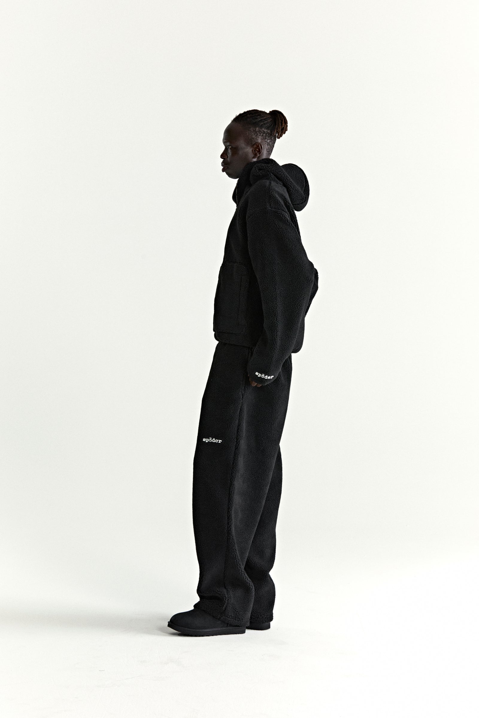 Black teddy fleece zip-up jacket and matching pants with white logo patches, worn by a person in profile.