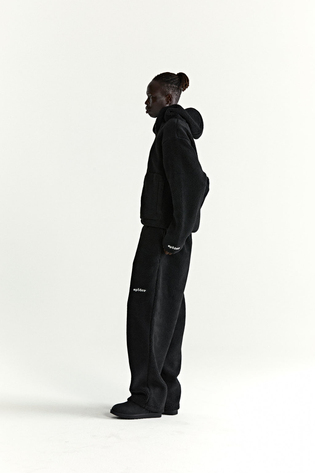 Black teddy fleece zip-up jacket and matching pants with white logo patches, worn by a person in profile.