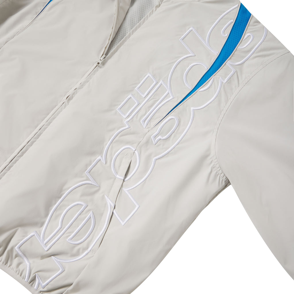 Beige nylon track jacket with white embroidered graphic text, full zip closure, and blue contrast panel detailing.