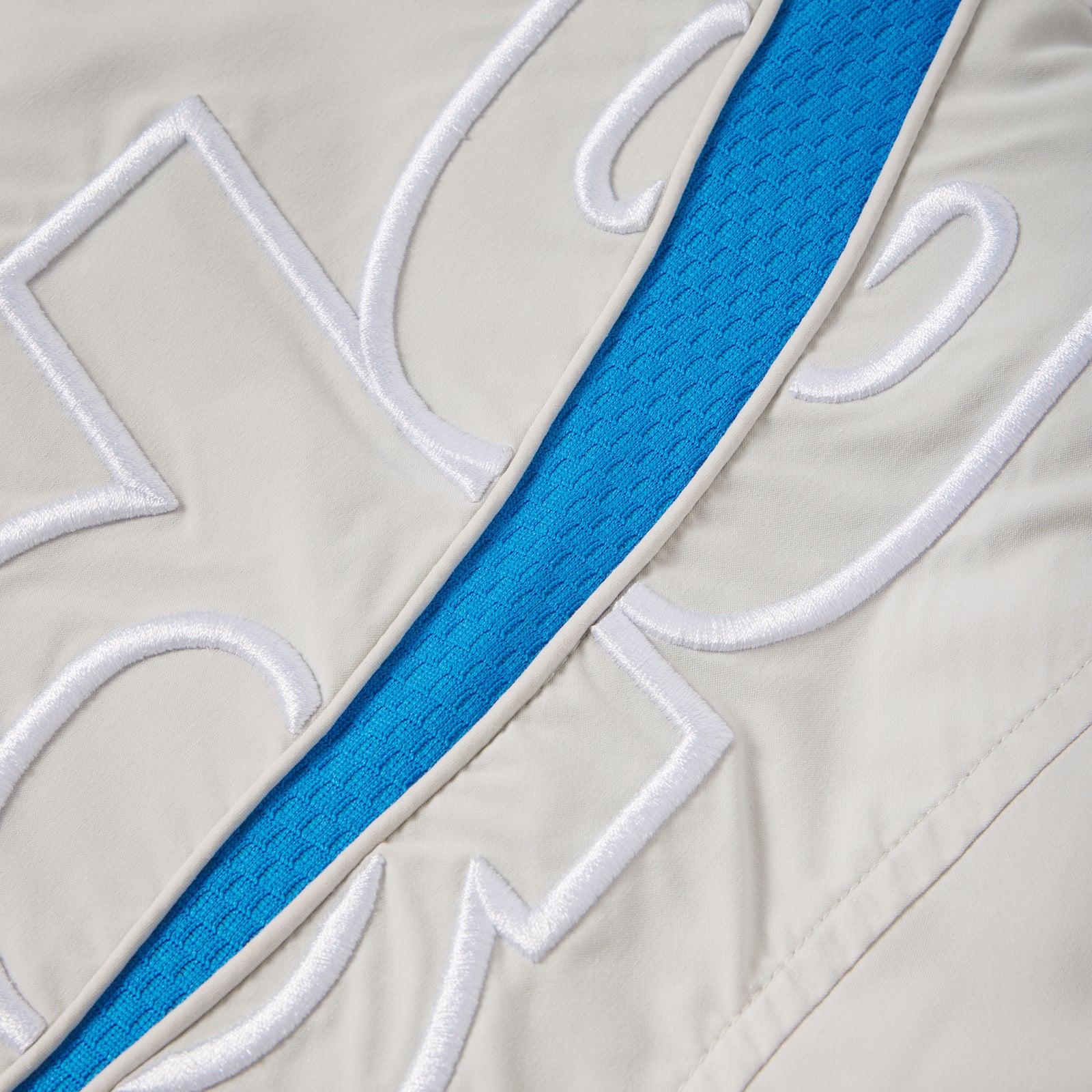 Beige nylon track jacket featuring a bright blue mesh contrast panel and white embroidered graphic design details.