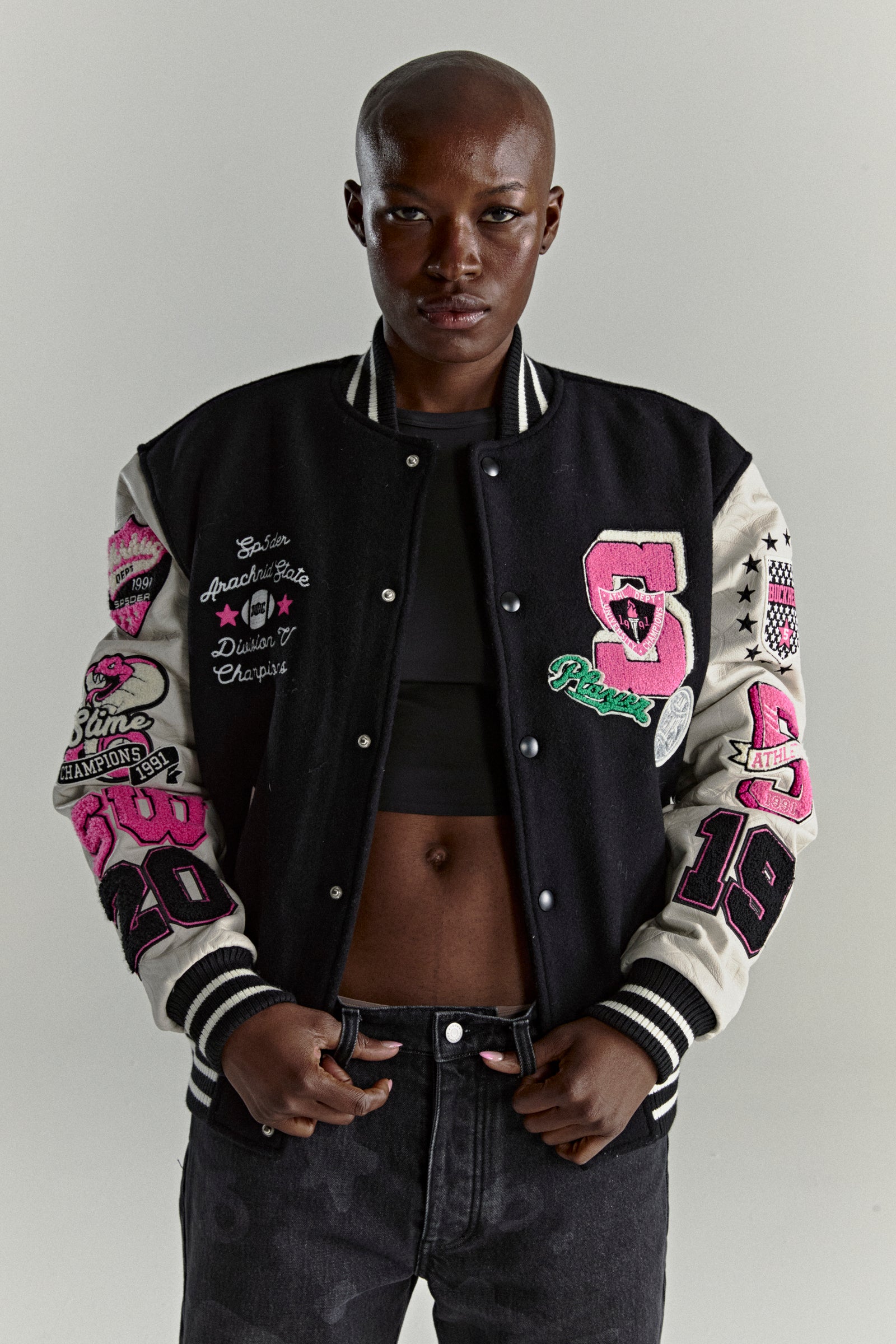 Black wool varsity jacket with contrasting leather sleeves, debossed web design, and multiple pink embroidered patches.