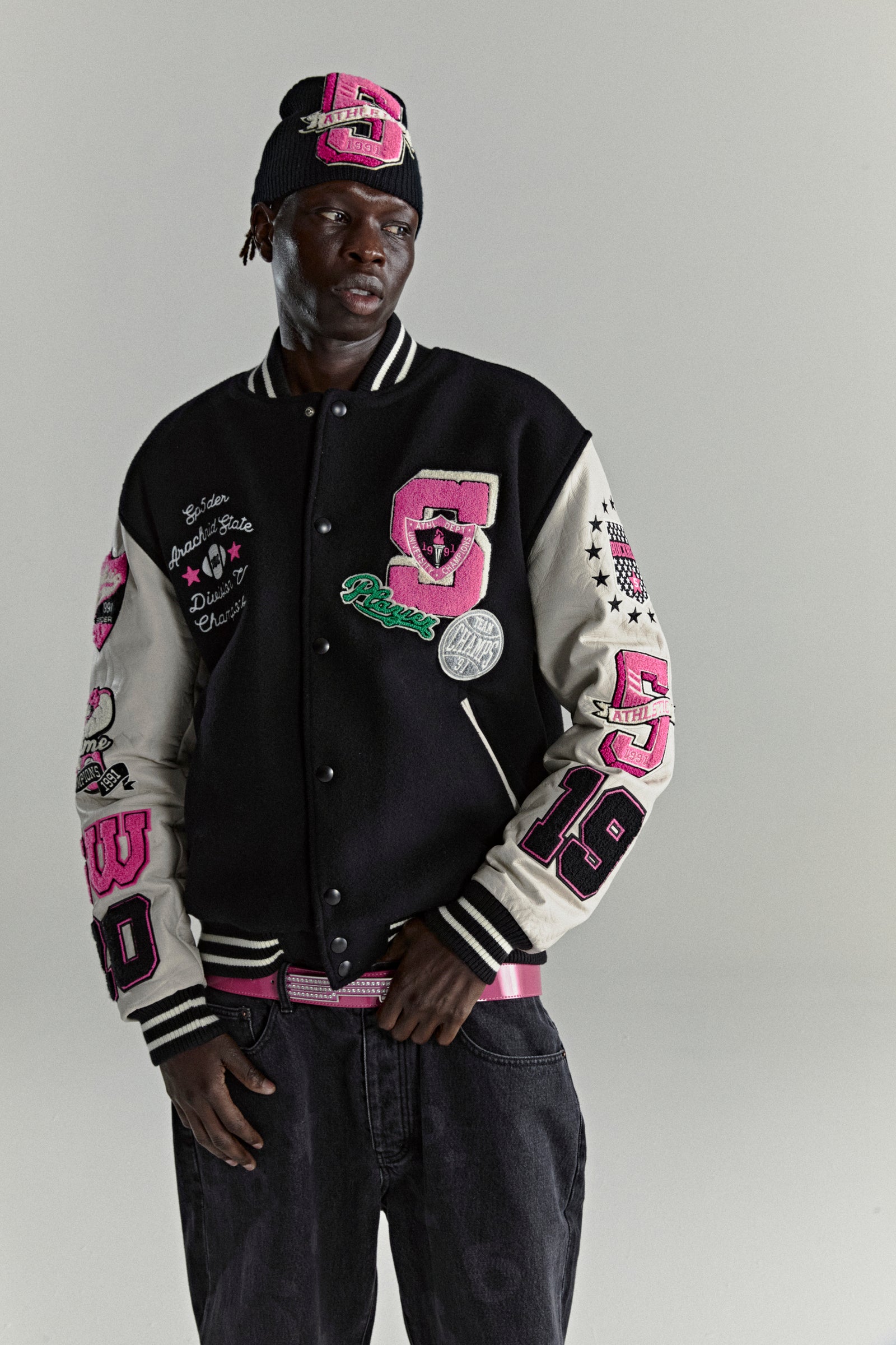 Black wool varsity jacket with cream leather sleeves, pink and white patches, and striped trim.