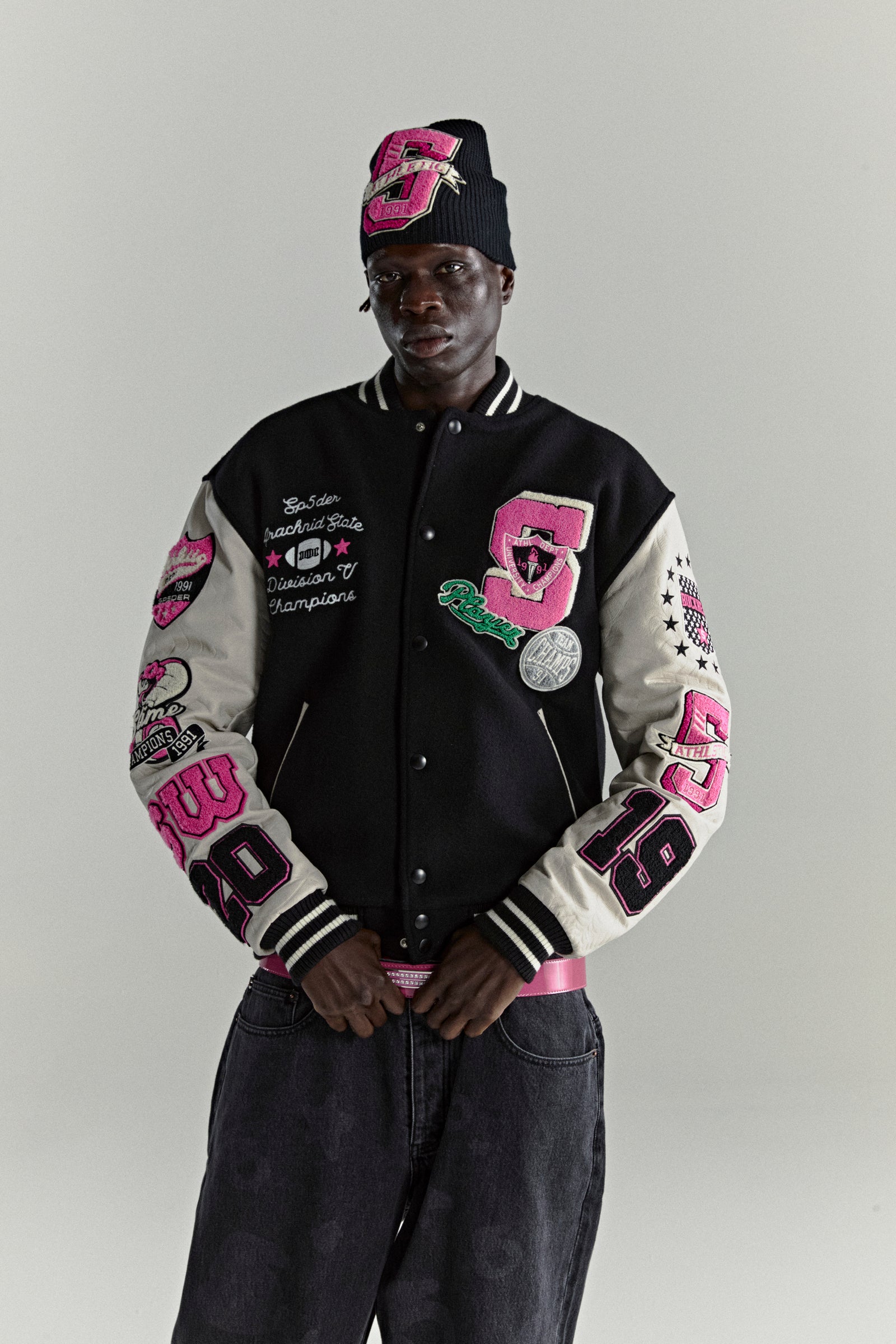 Black wool varsity jacket with contrasting leather sleeves, multiple pink embroidered patches, and striped ribbed trim.