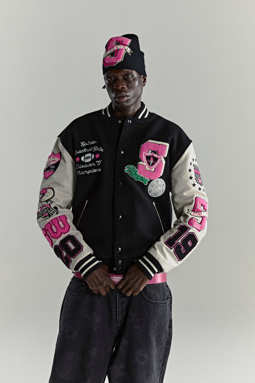 Black wool varsity jacket with contrasting leather sleeves, multiple pink embroidered patches, and striped ribbed trim.