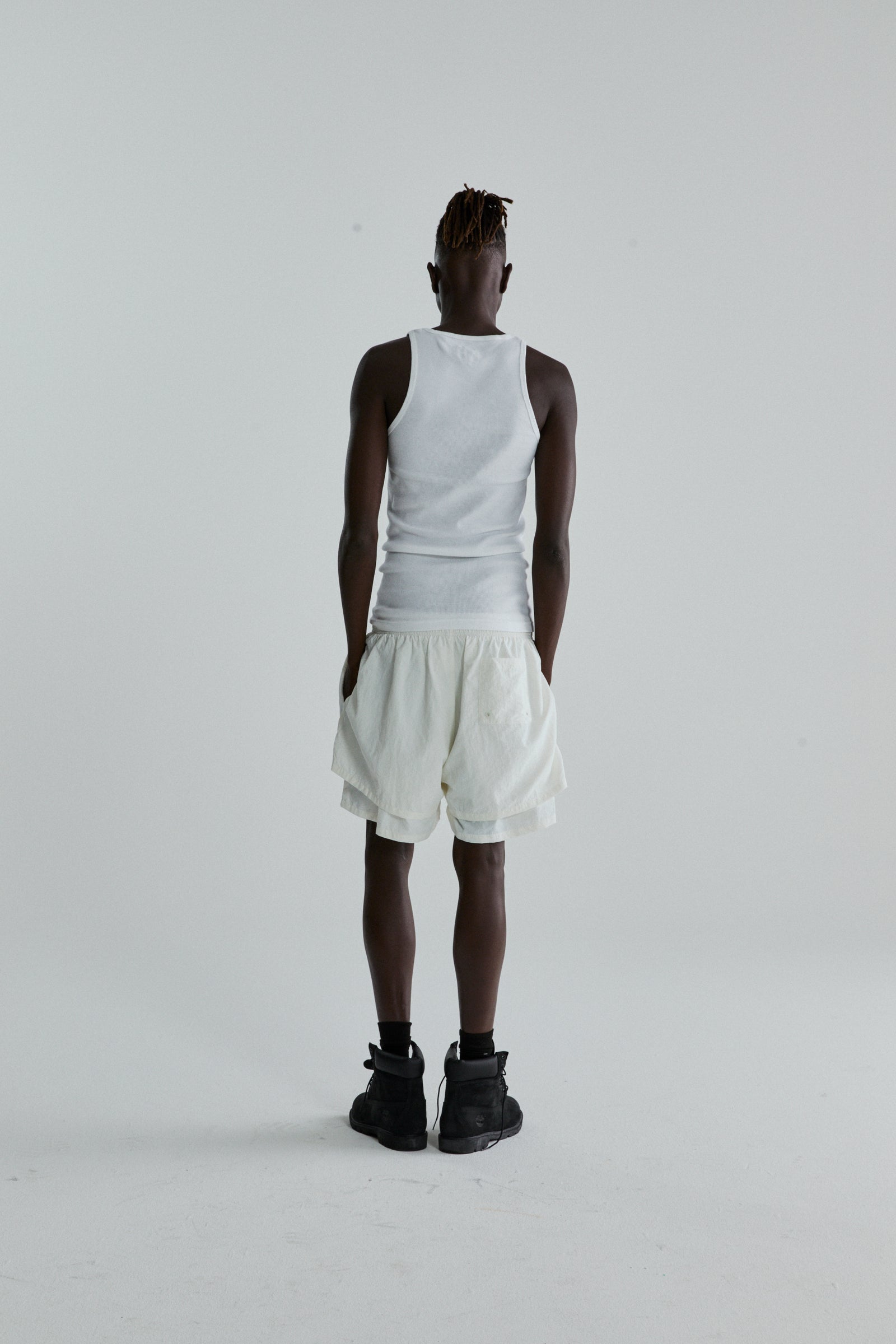 Rear view of a person wearing white OG WEB DOUBLE LAYER SHORT with a high-low design and a white tank top.