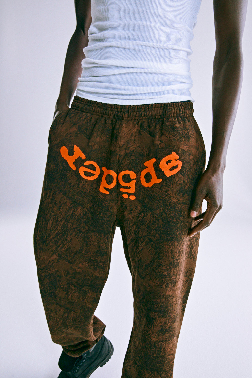 Brown tree camo sweatpants with orange "SP5DER" text, drawstring waist, and elasticized cuffs.