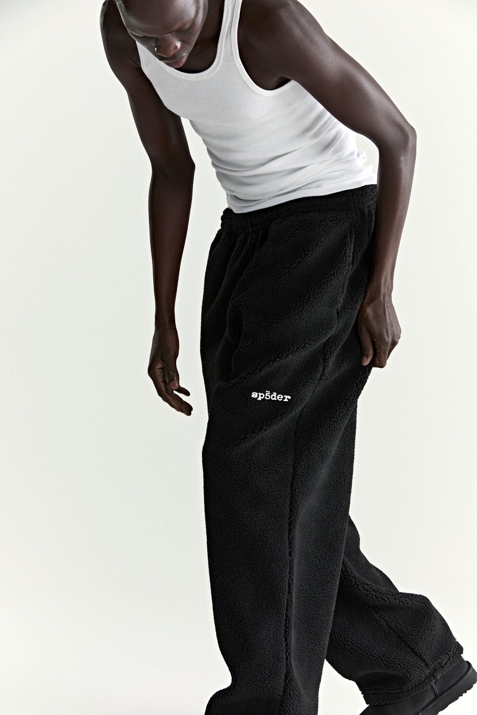 Black teddy fleece pant with white "sp5der" logo on leg, worn by a person in a white tank top.