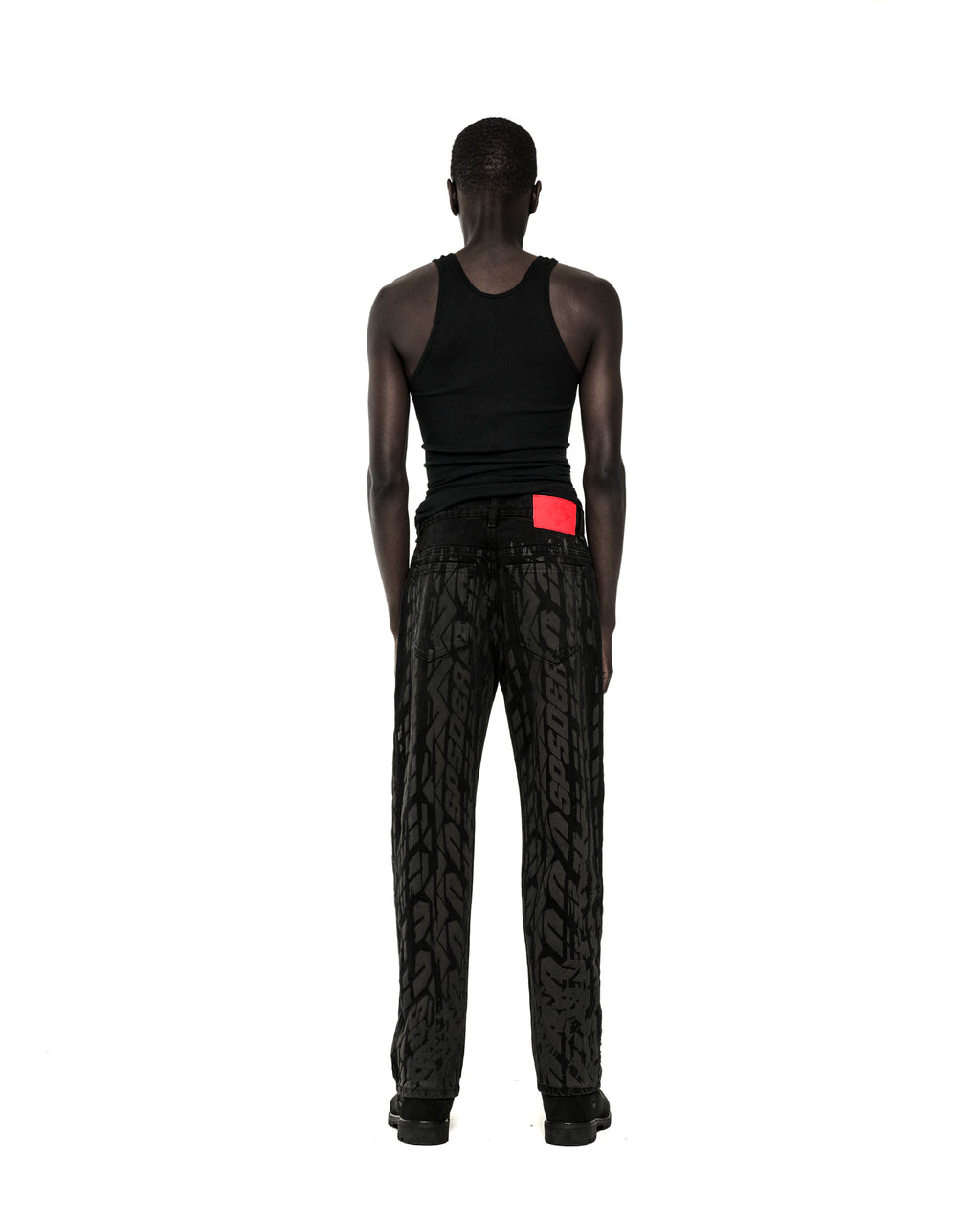 Man wearing black Burnout Denim pants with all-over tire track design and a red branded back patch.