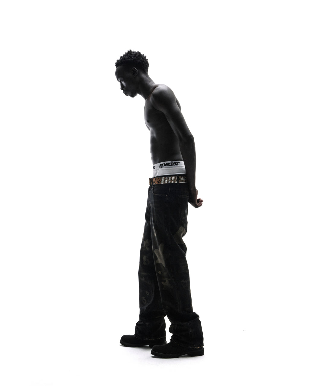 A shirtless model in dark patterned jeans and a studded belt stands in profile against a bright white background.