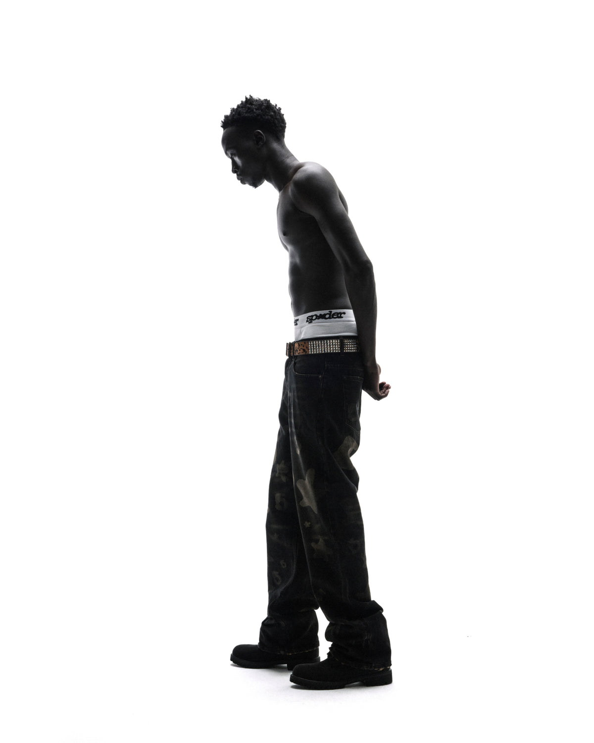 A shirtless model in dark patterned jeans and a studded belt stands in profile against a bright white background.