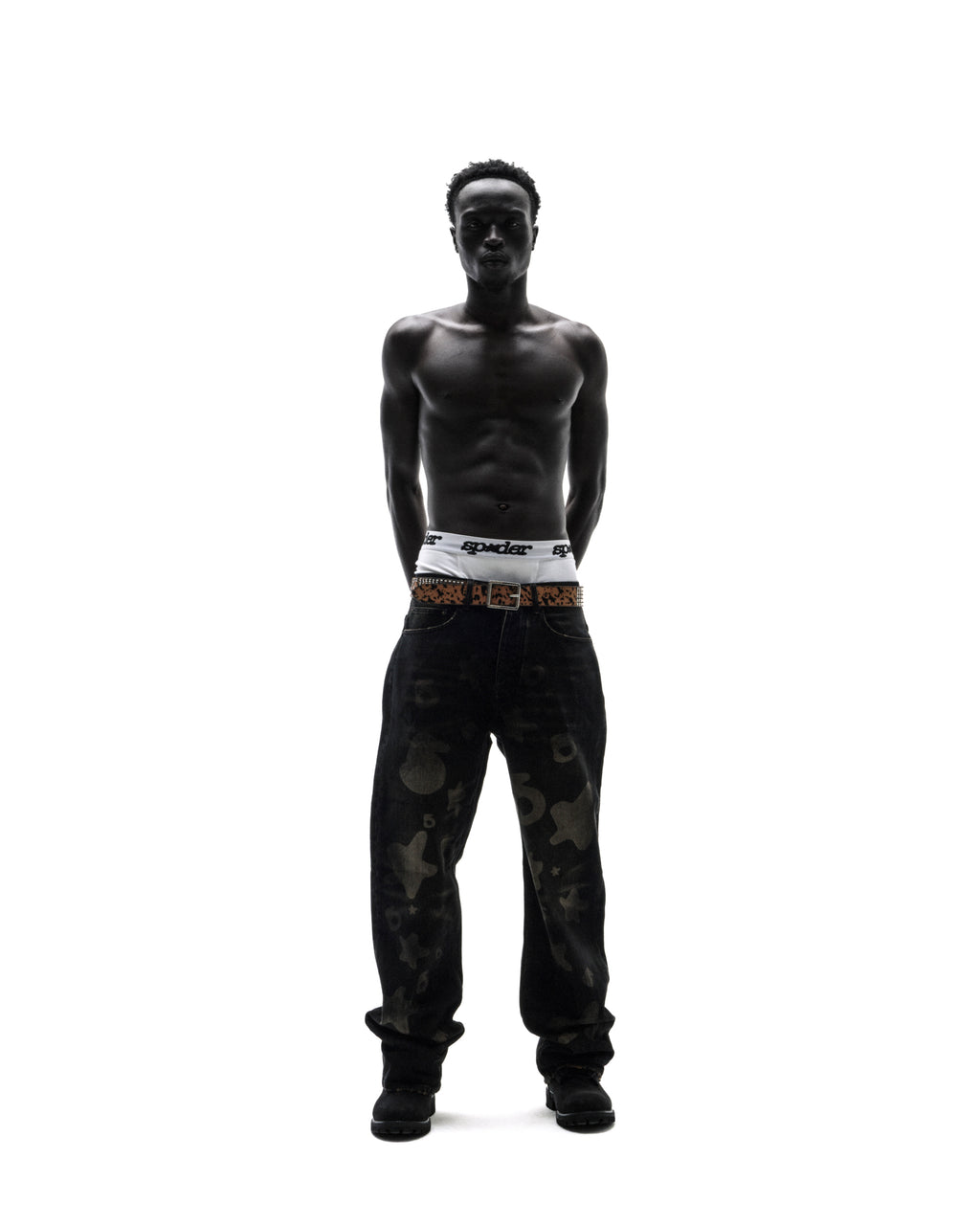 Shirtless model wearing white Sp5der briefs, a leopard print belt, and baggy black patterned jeans against white.