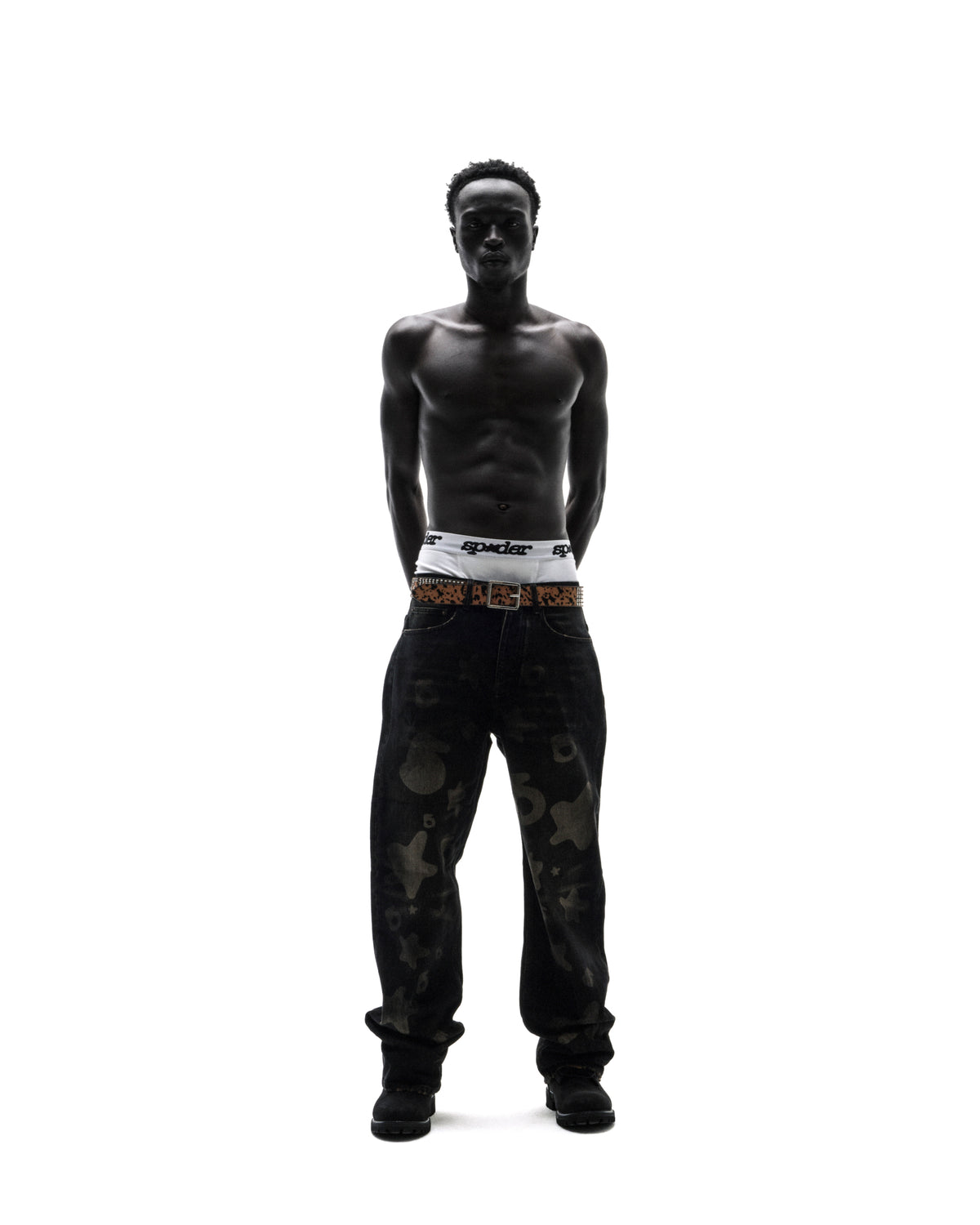 Shirtless model wearing white Sp5der briefs, a leopard print belt, and baggy black patterned jeans against white.