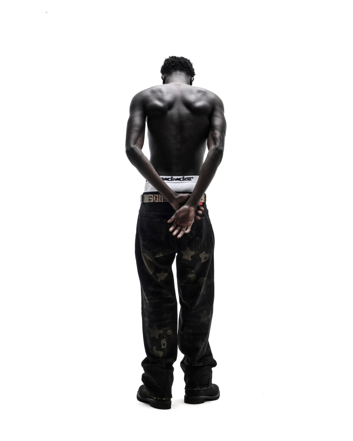 Shirtless model seen from behind, wearing black patterned jeans, a studded belt, and white boxers against a white background.