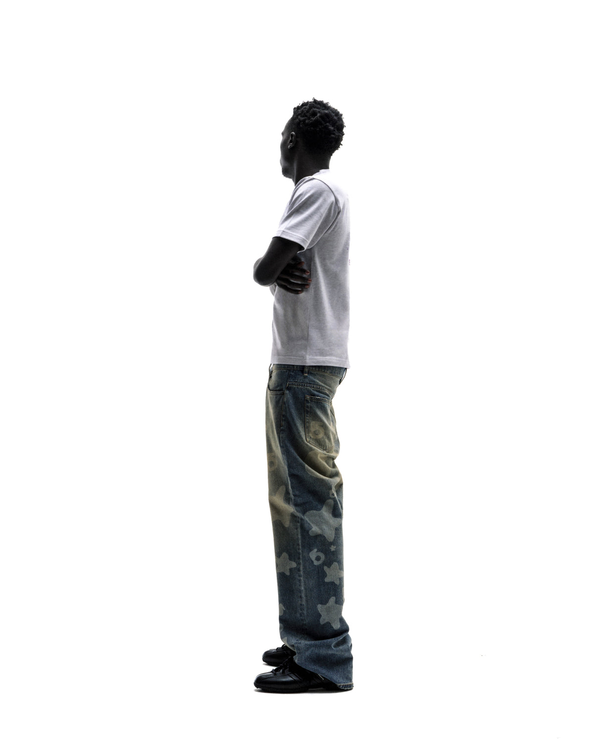 Model in a white t-shirt and baggy blue jeans with star patterns, standing with arms crossed against a white background.