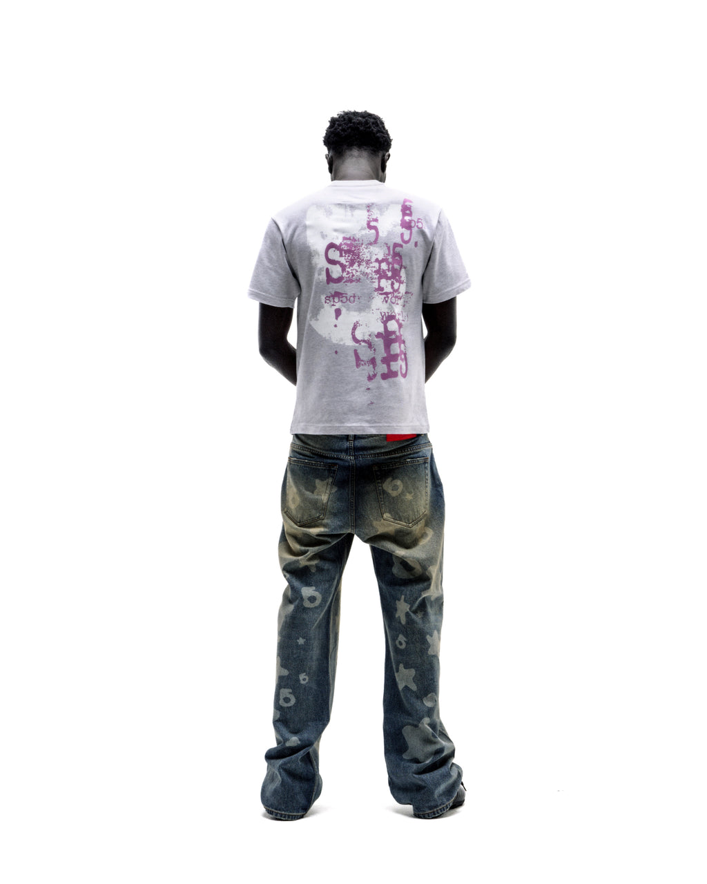 A model seen from behind wearing a grey t-shirt with purple graphic prints and distressed blue jeans with star patterns.