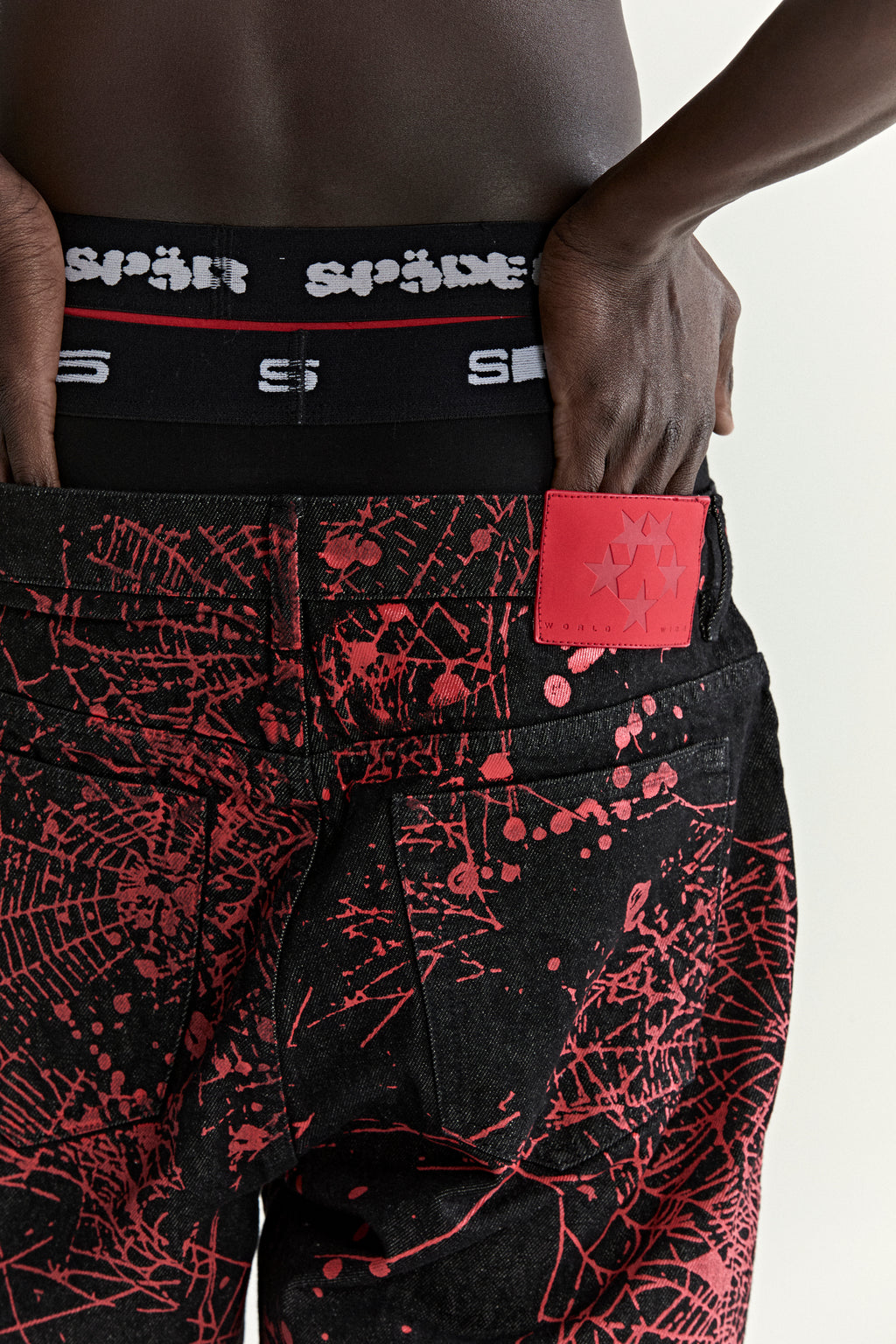 Black denim pants with all-over red web print, five-pocket styling, and a red leather branded patch.
