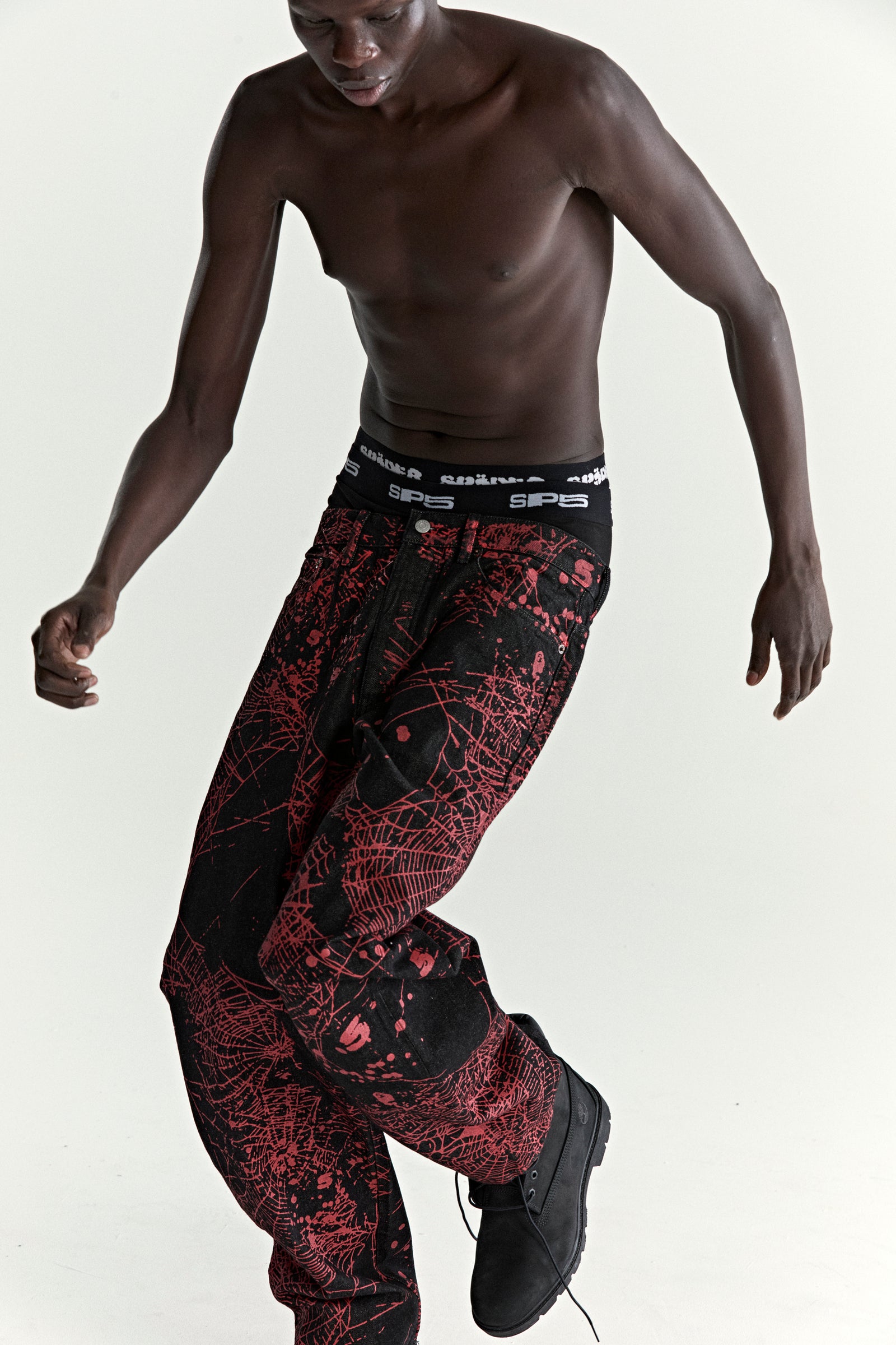 Black denim pants with red all-over web print, classic straight leg fit, and SP5DER branding.