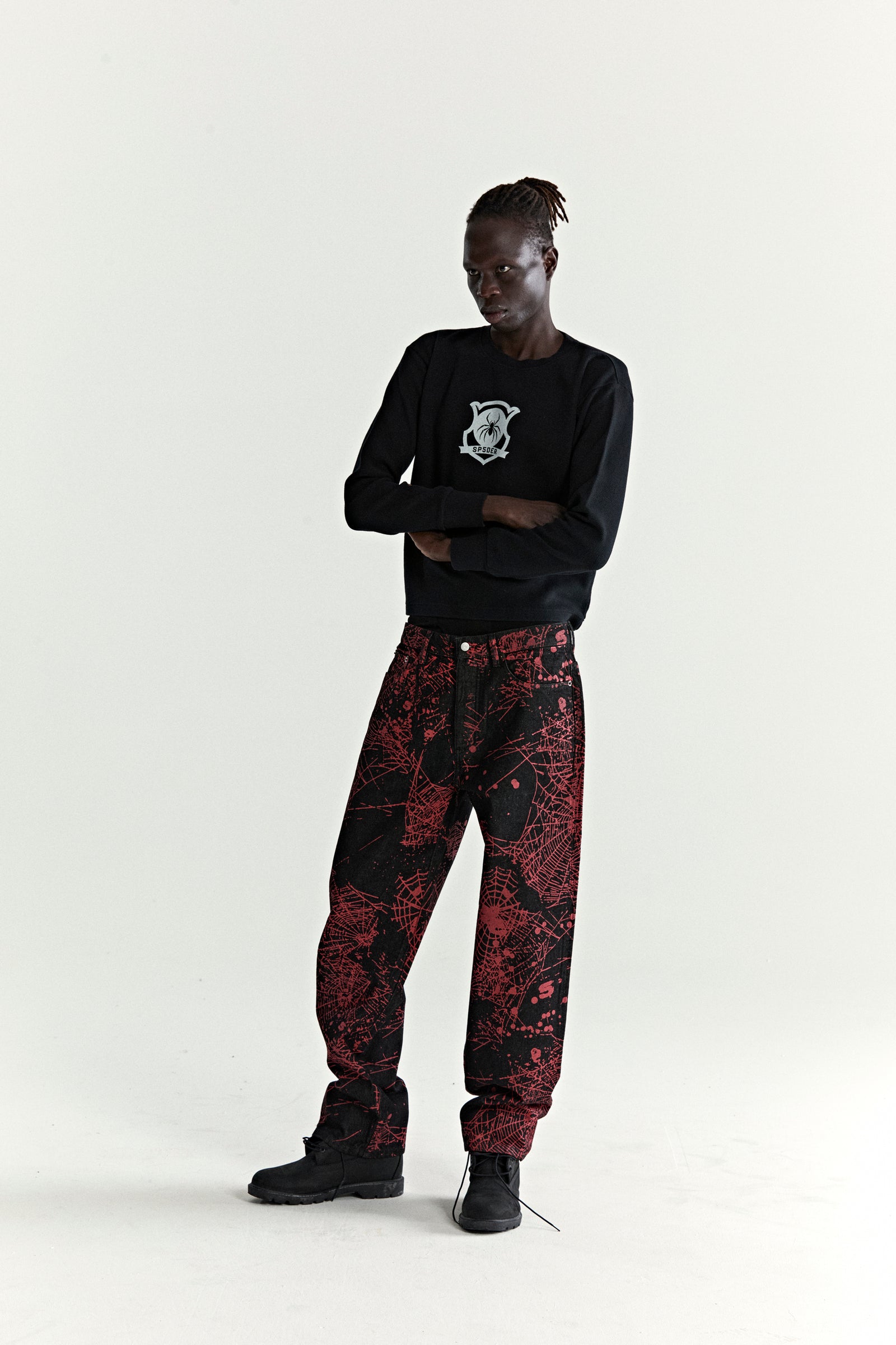 Model wearing V2 WEB DENIM pants: black denim with red all-over web print, straight leg fit.