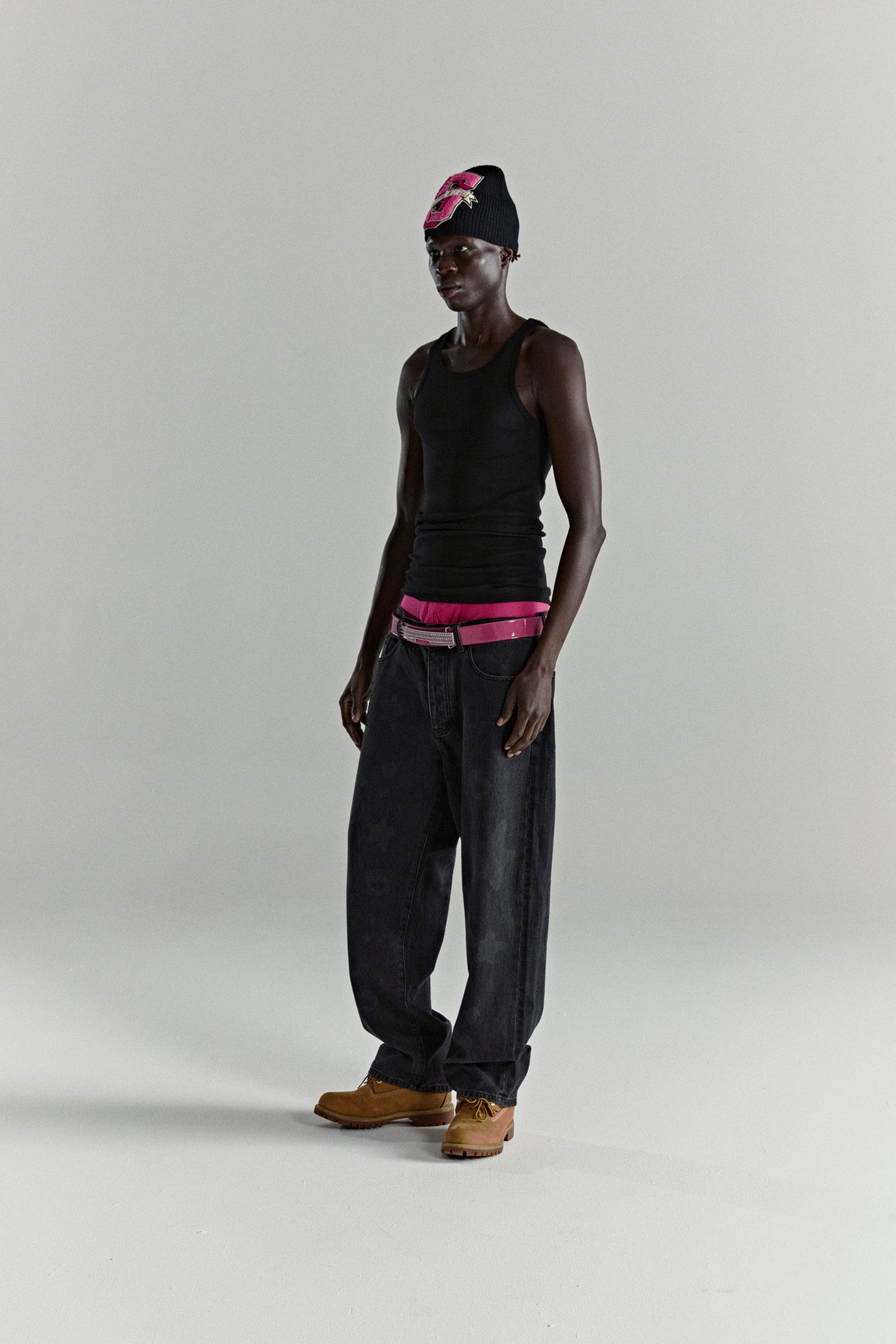 Model wearing faded wash Beluga denim pants, black tank top, pink belt, and floral beanie.
