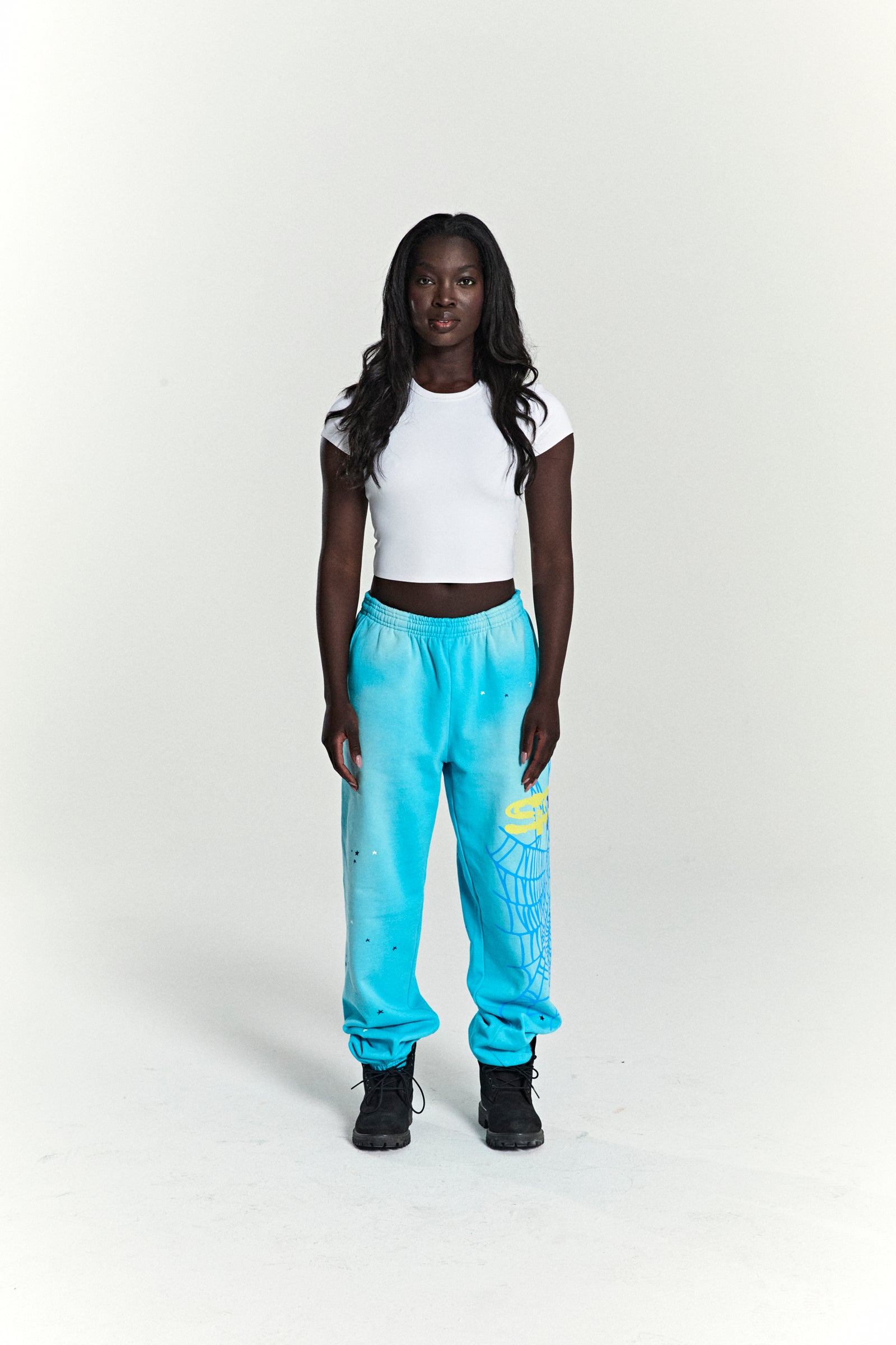 Woman wearing blue HEAVY WEB SWEATPANTS with SP5DER web graphic, scattered stars, and elasticized cuffs.